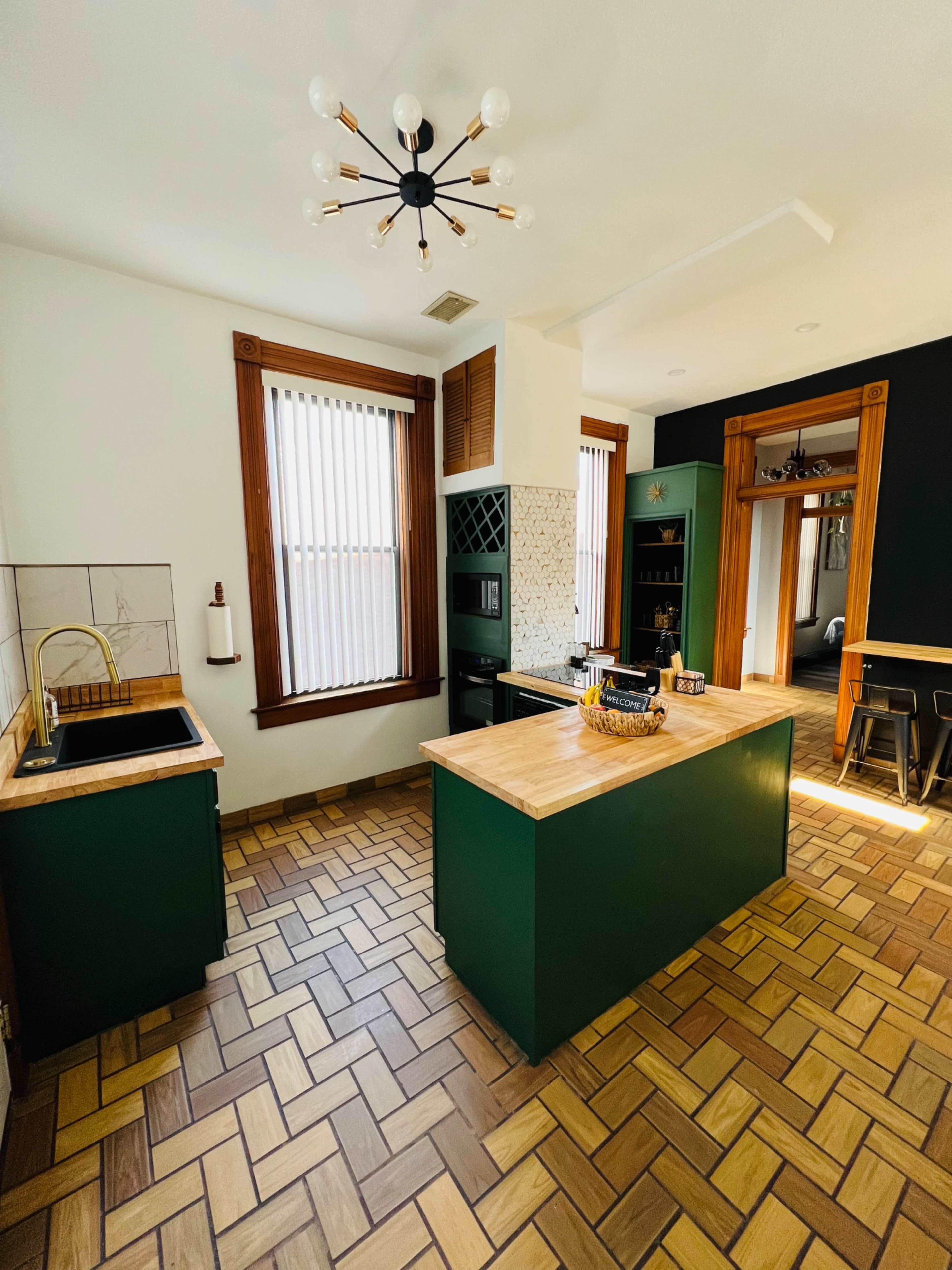 A well-lit kitchen featuring green cabinetry, a wooden island, and brown tiled flooring with a geometric pattern.
