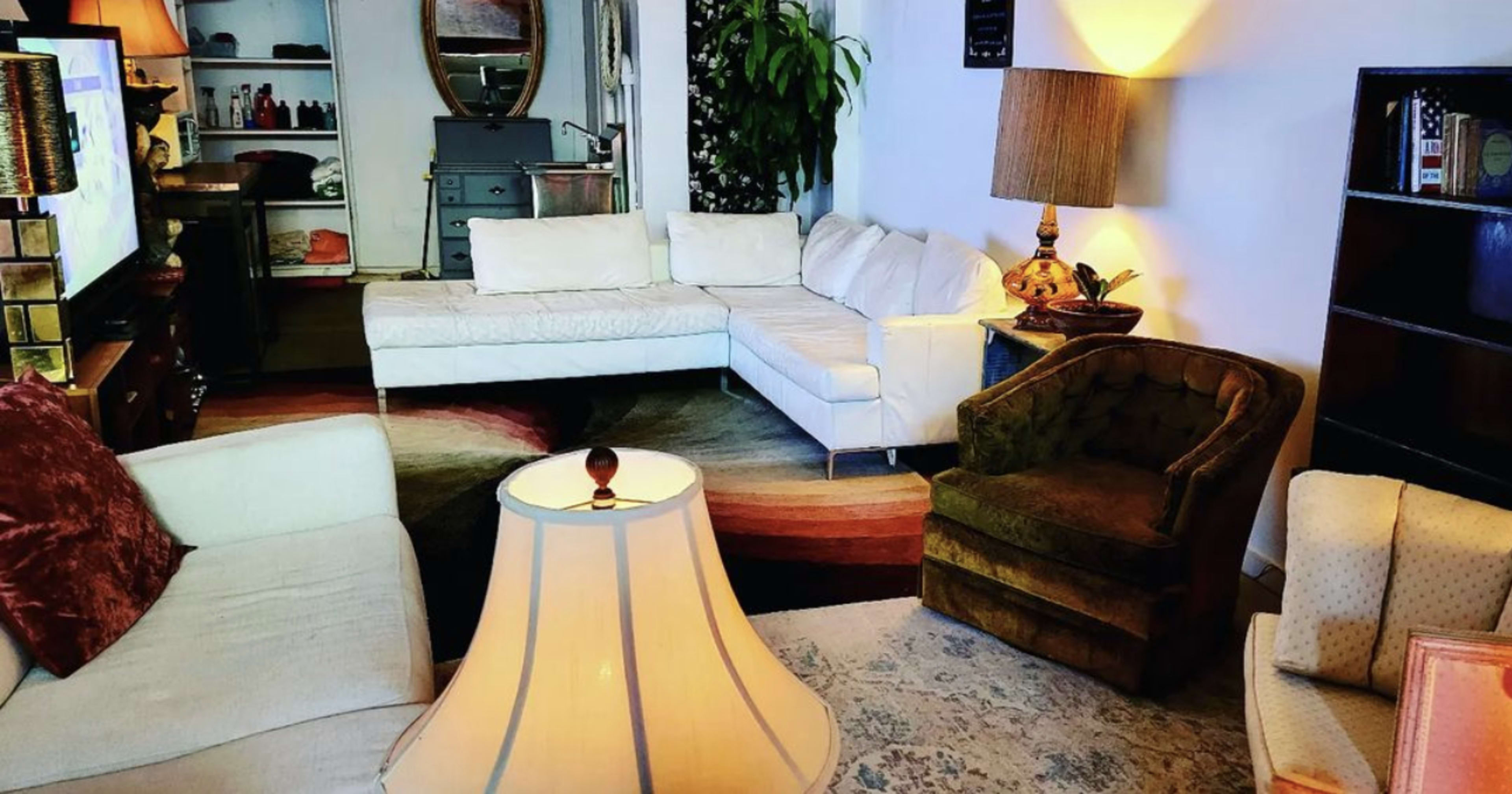 A cozy living room with white sectional sofas, a green armchair, a table lamp, and a patterned rug, along with a small kitchenette visible in the background.
