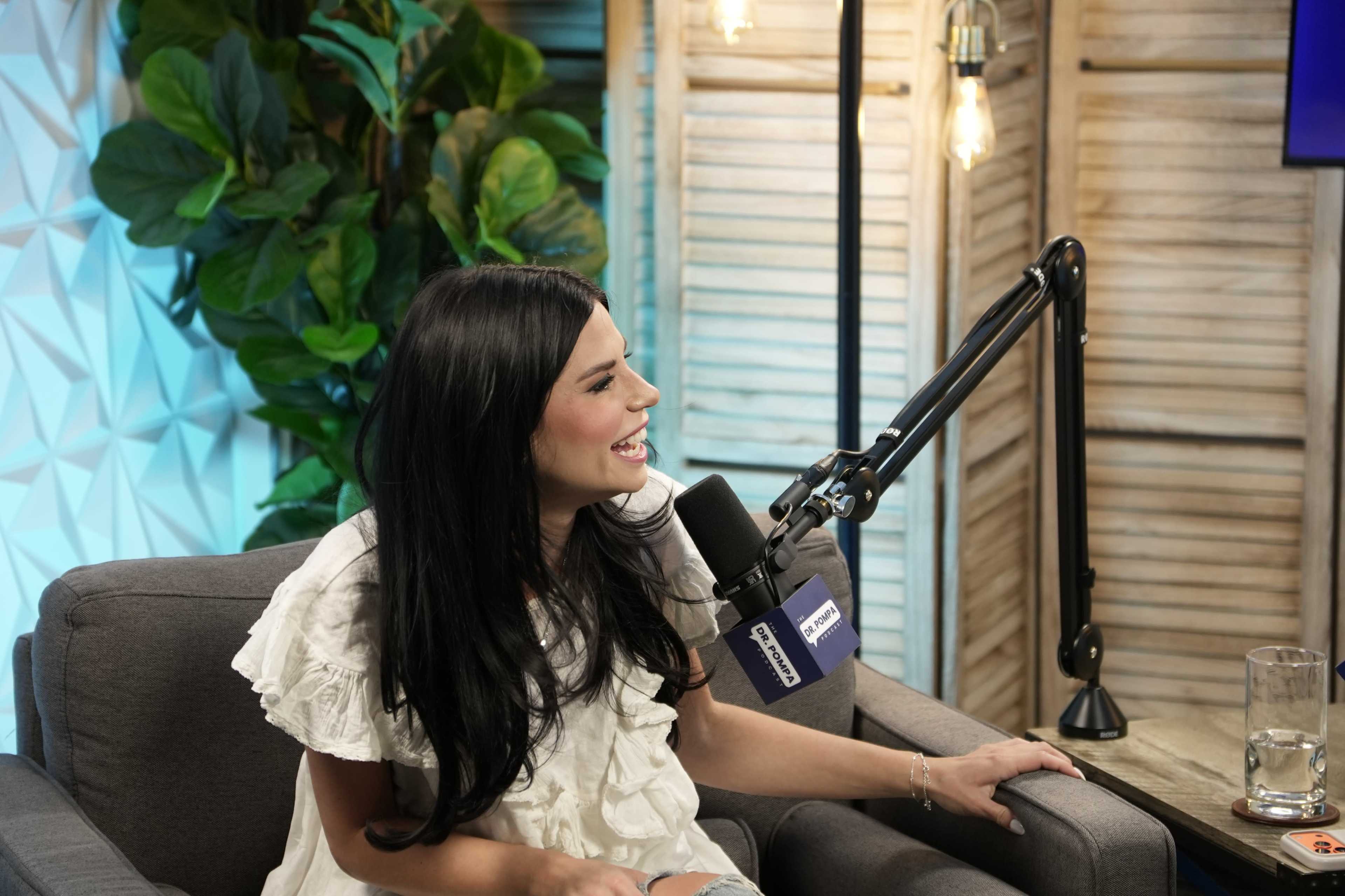A woman with long black hair is sitting on a gray couch, smiling while speaking into a microphone in a well-lit studio setting.