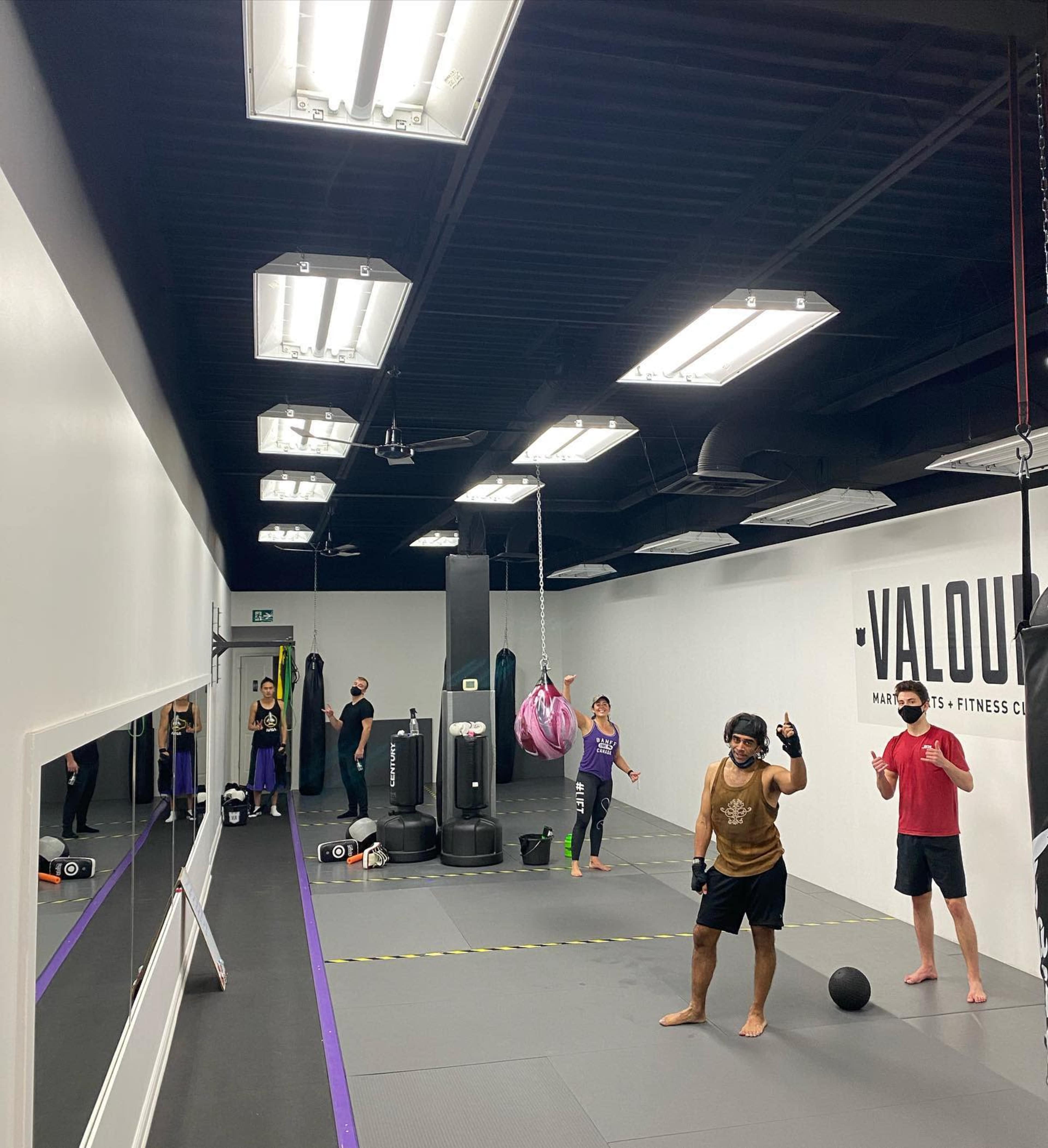 A group of people are engaged in various fitness activities inside a gym, featuring workout equipment and a boxing bag in a mirrored space.