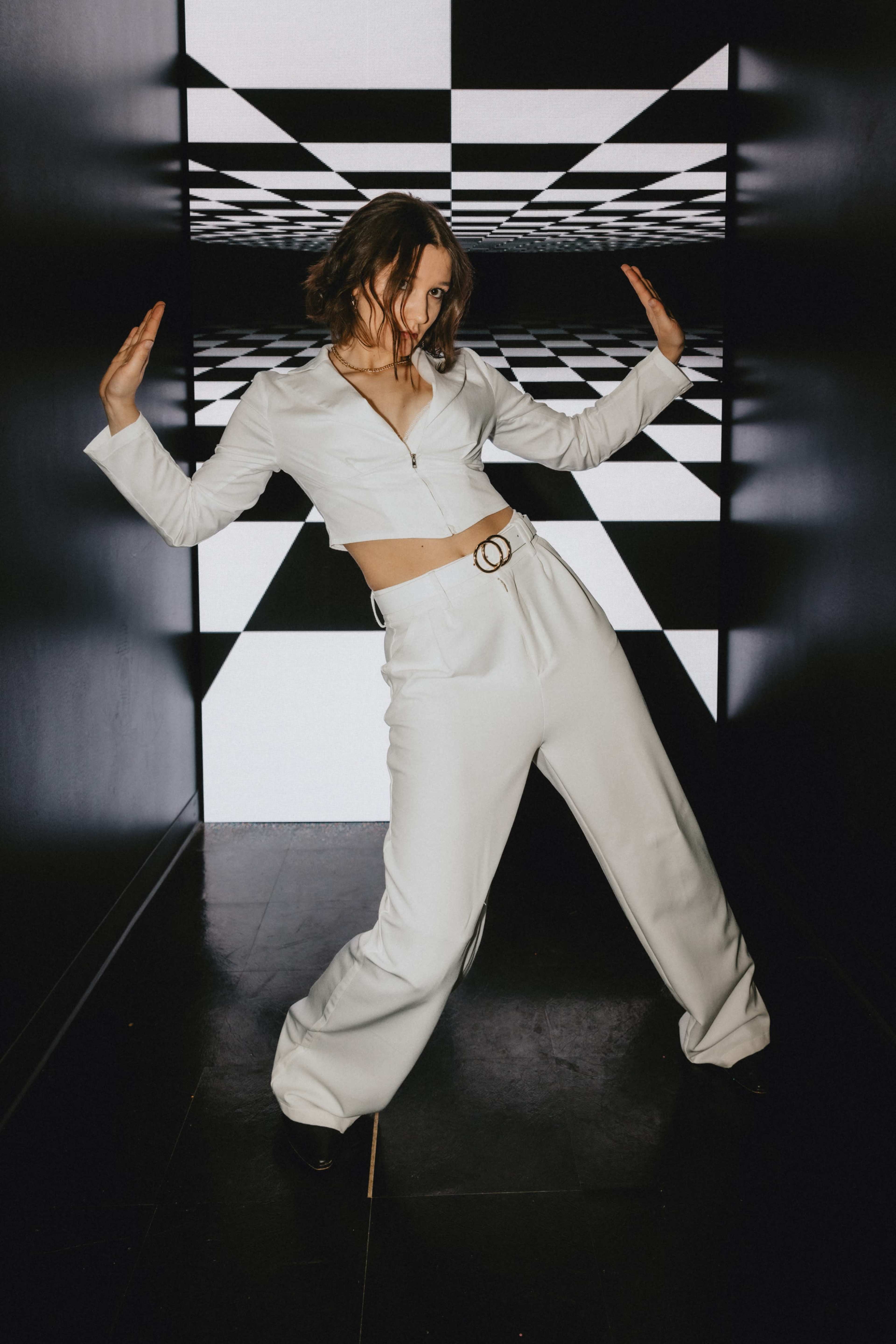 A model poses in a white outfit against a backdrop of alternating black and white geometric patterns.