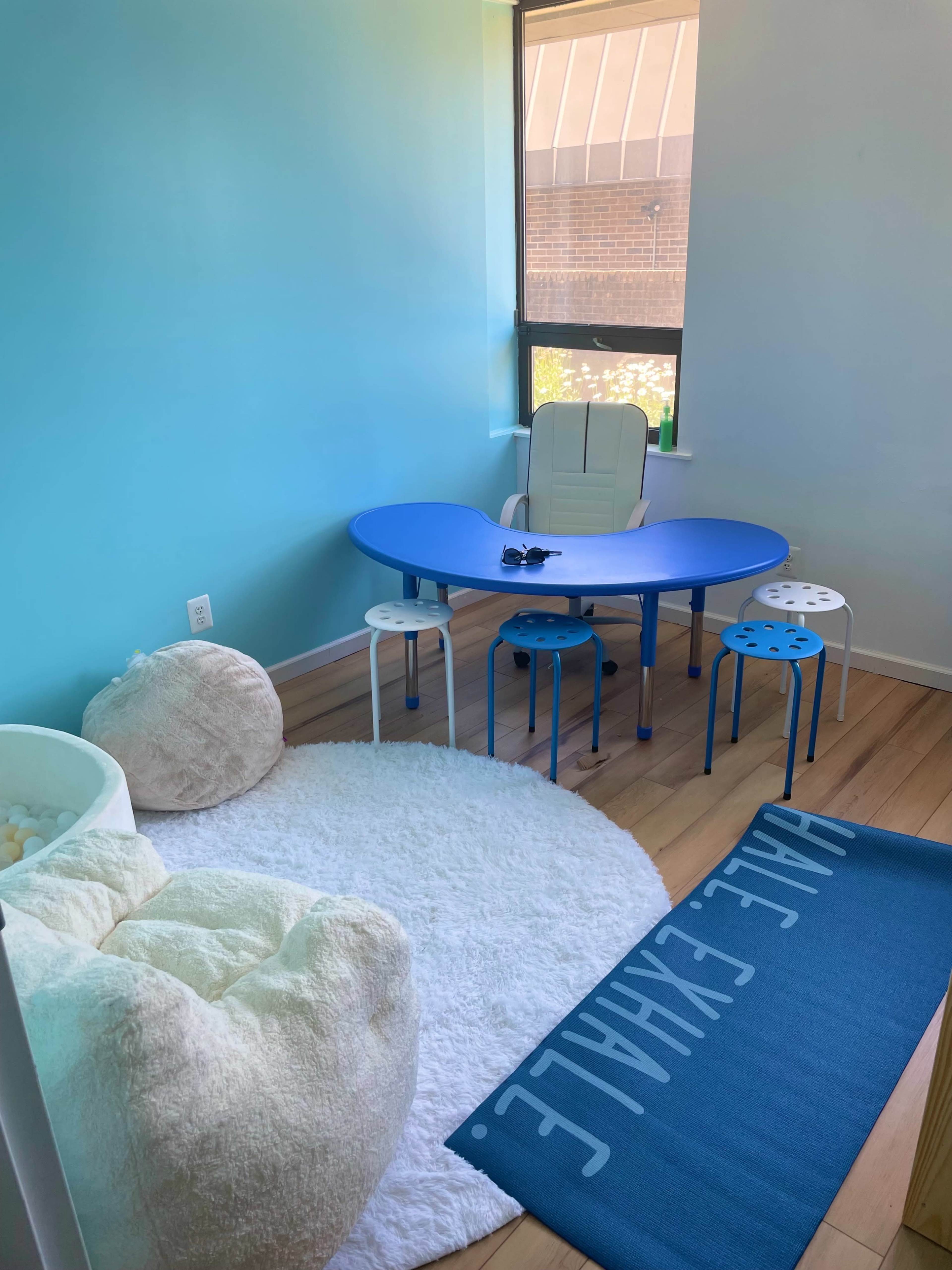 A small room features a blue tabletop surrounded by chairs and small stools, a white rug, and a cozy bean bag chair.