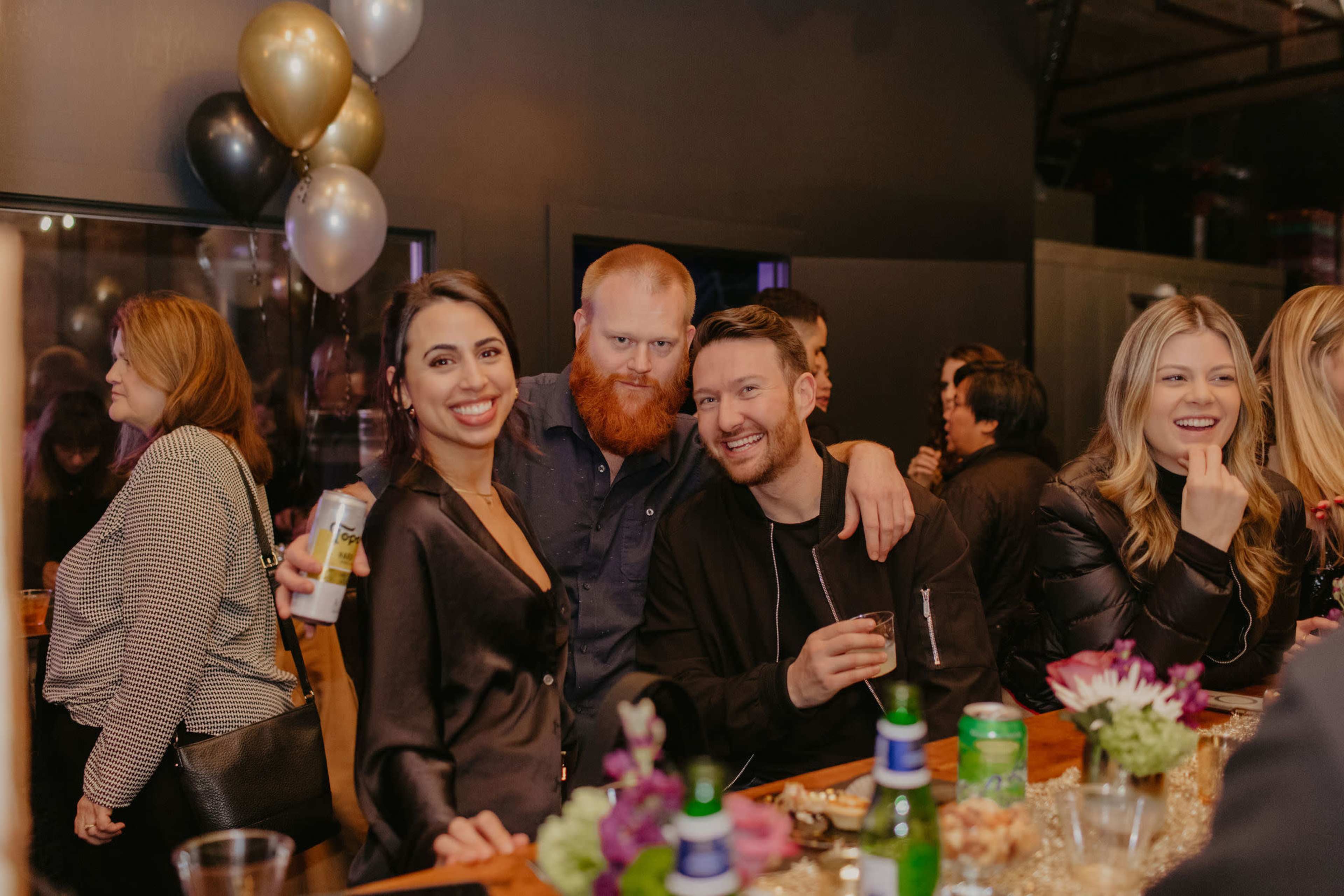 A group of four people poses together at a bar during a social gathering, with drinks and decorations in the background.