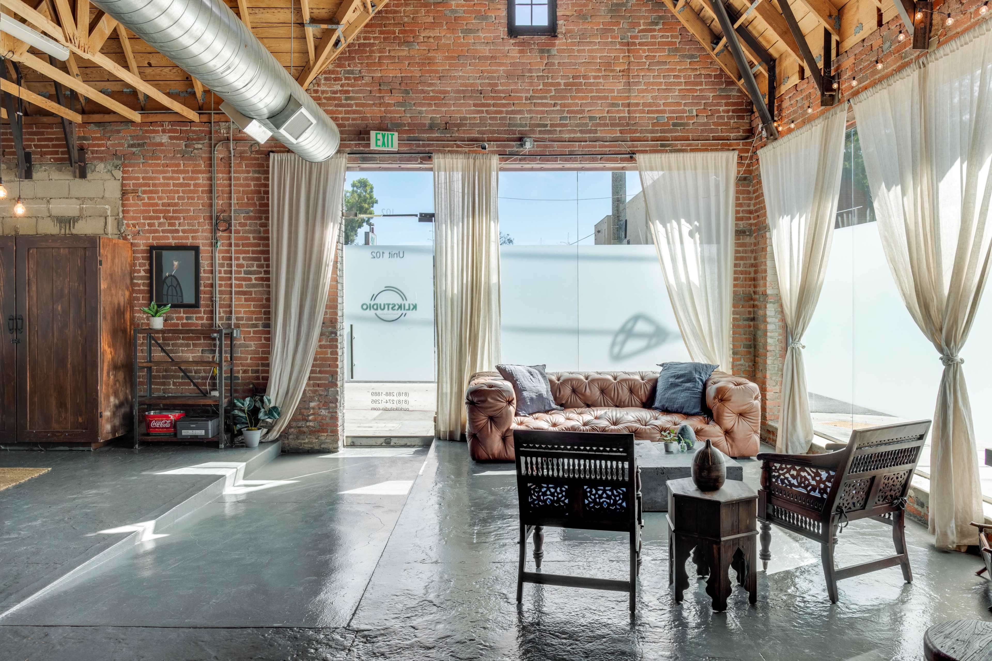 A spacious, modern interior with exposed brick walls, large windows, a brown leather sofa, and wooden furniture.