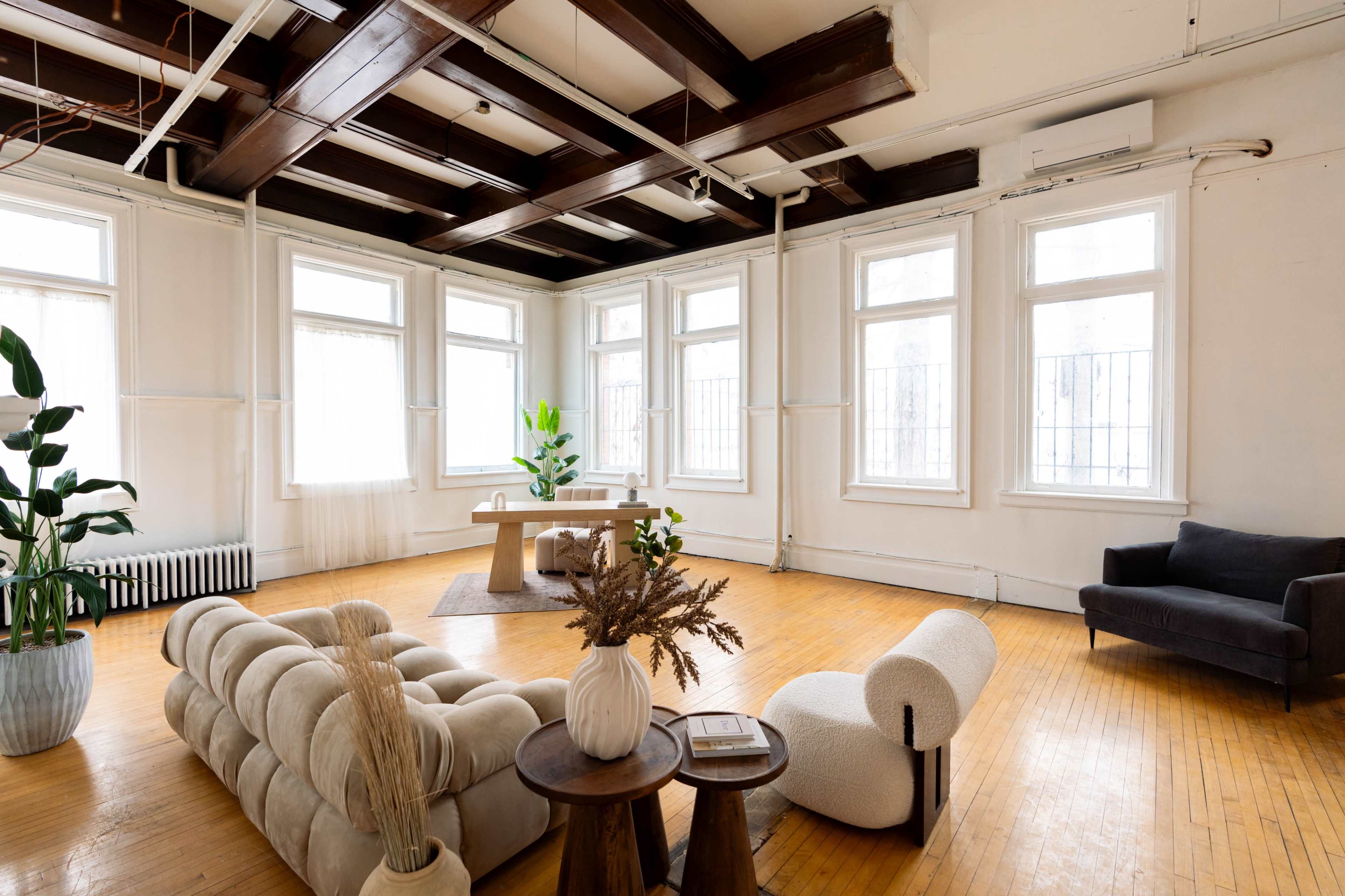 The image shows a spacious interior with wooden flooring, large windows, and a minimalistic arrangement of furniture, including a couch, a chair, a coffee table, and decorative plants.