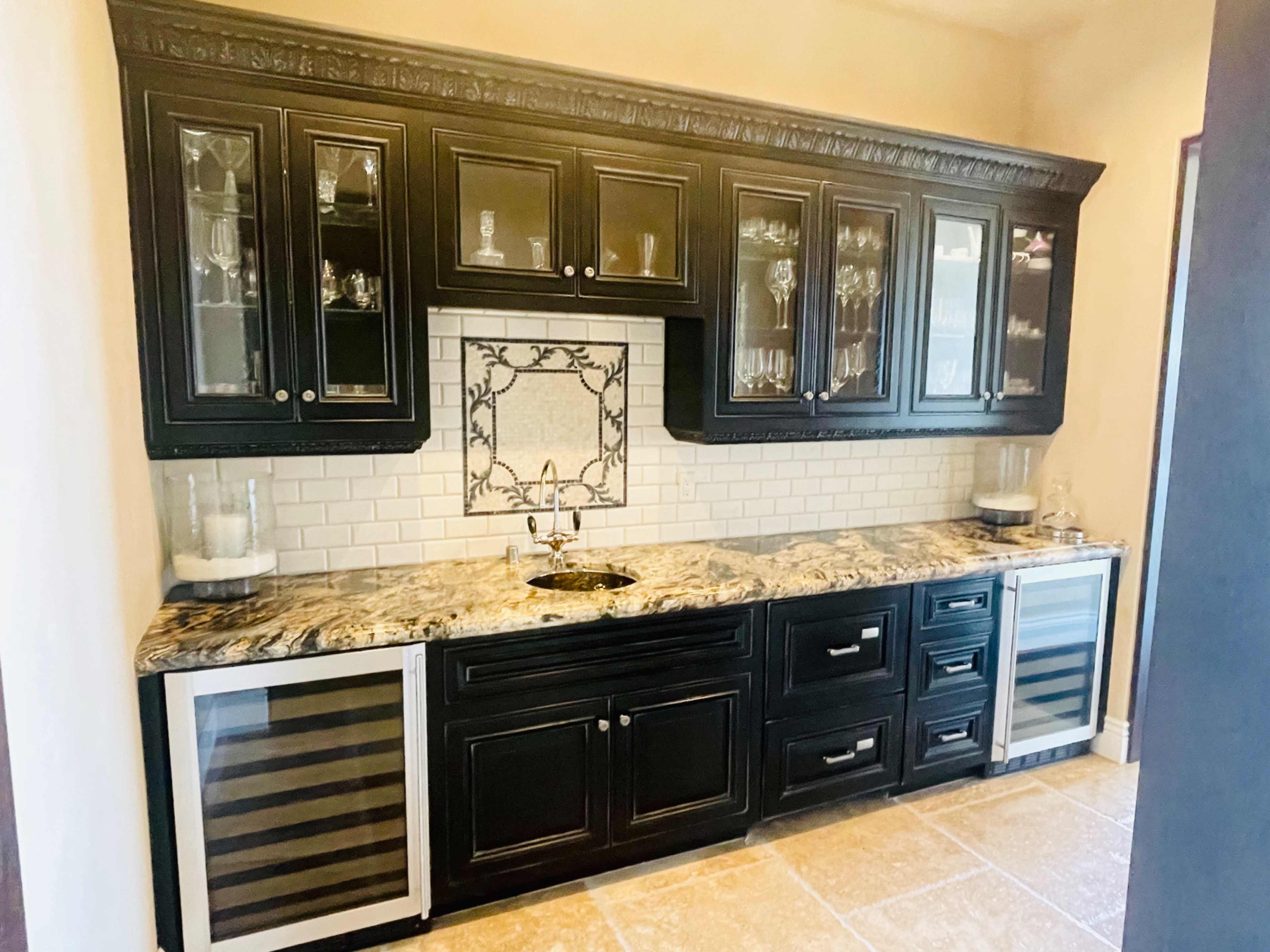 A kitchen bar area with black cabinets, a granite countertop, a backsplash featuring a decorative tile, and two wine coolers.