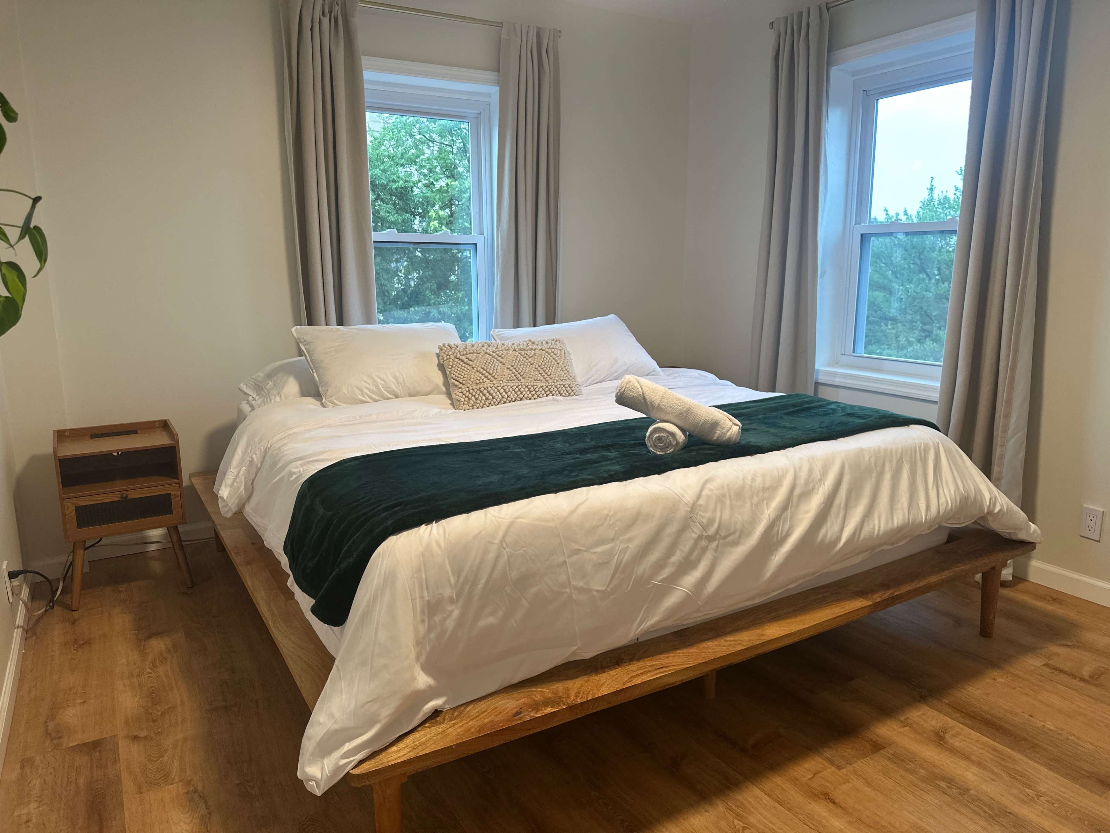 The image shows a bedroom featuring a large bed with a white comforter and green throw blanket, flanked by two windows with light curtains.