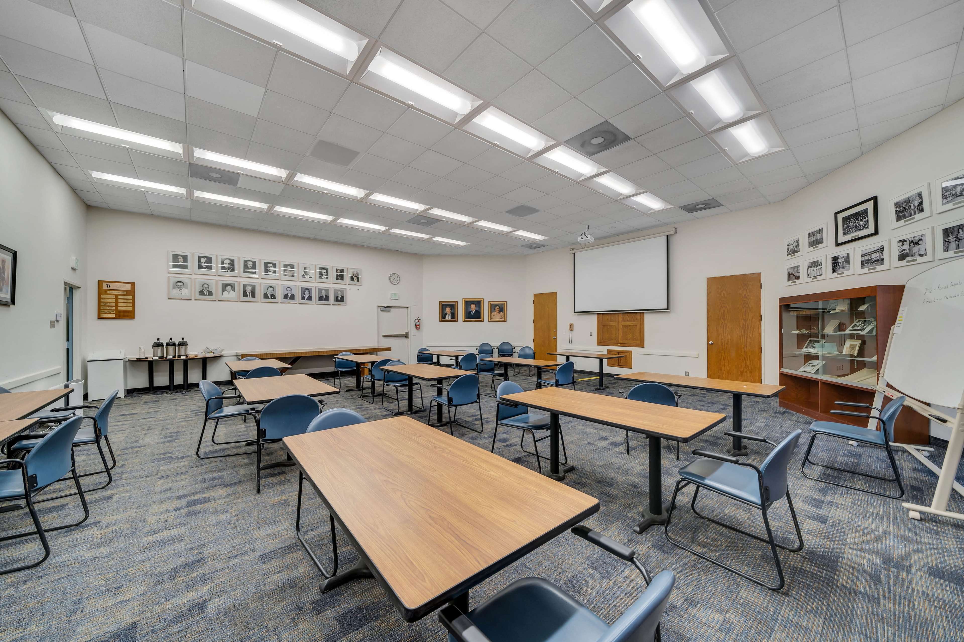 A spacious classroom features multiple tables and chairs arranged for a group setting, with framed photographs and a screen on the walls.