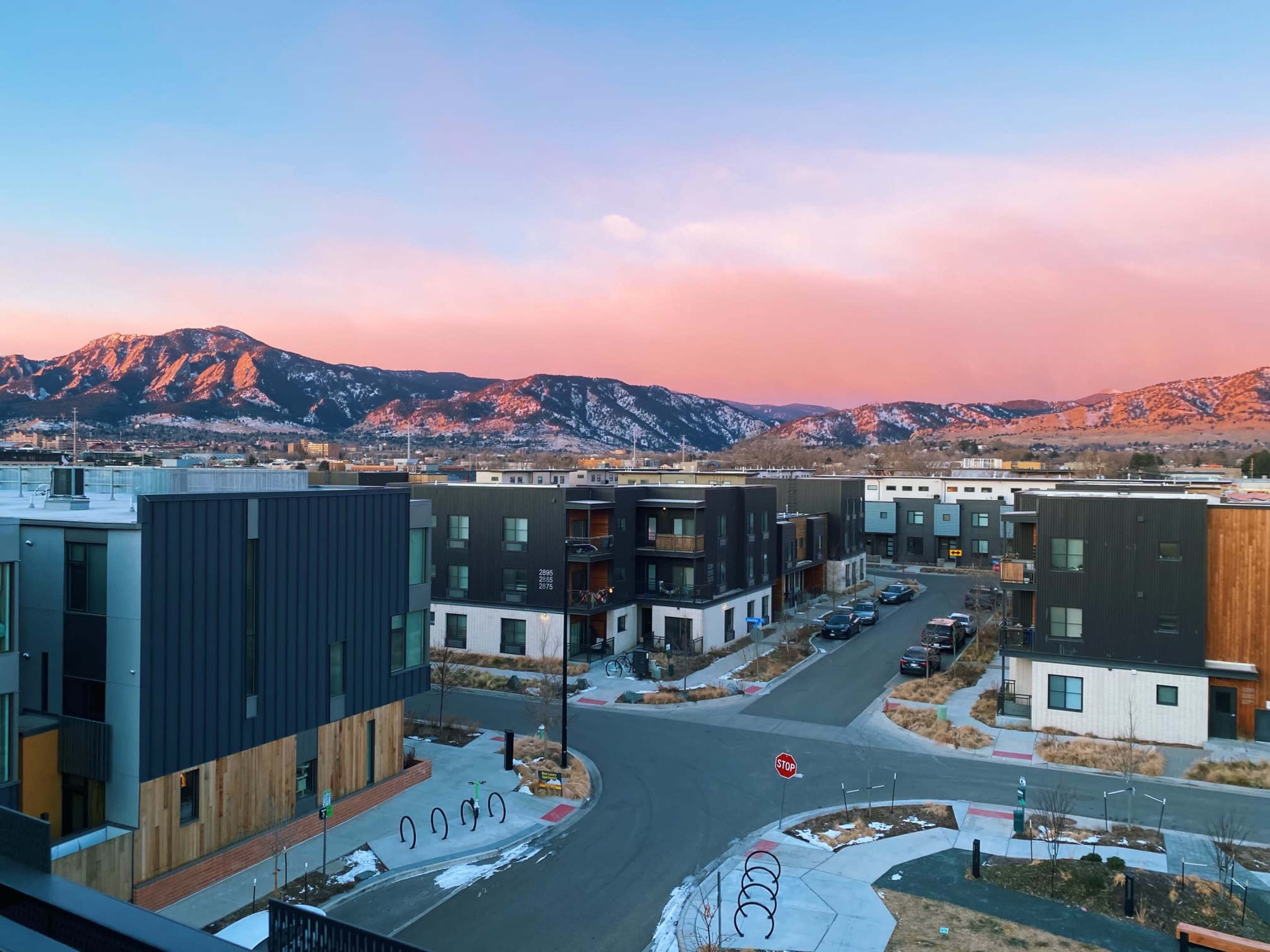 A suburban neighborhood with modern buildings and a backdrop of snow-capped mountains under a colorful sky at dusk.