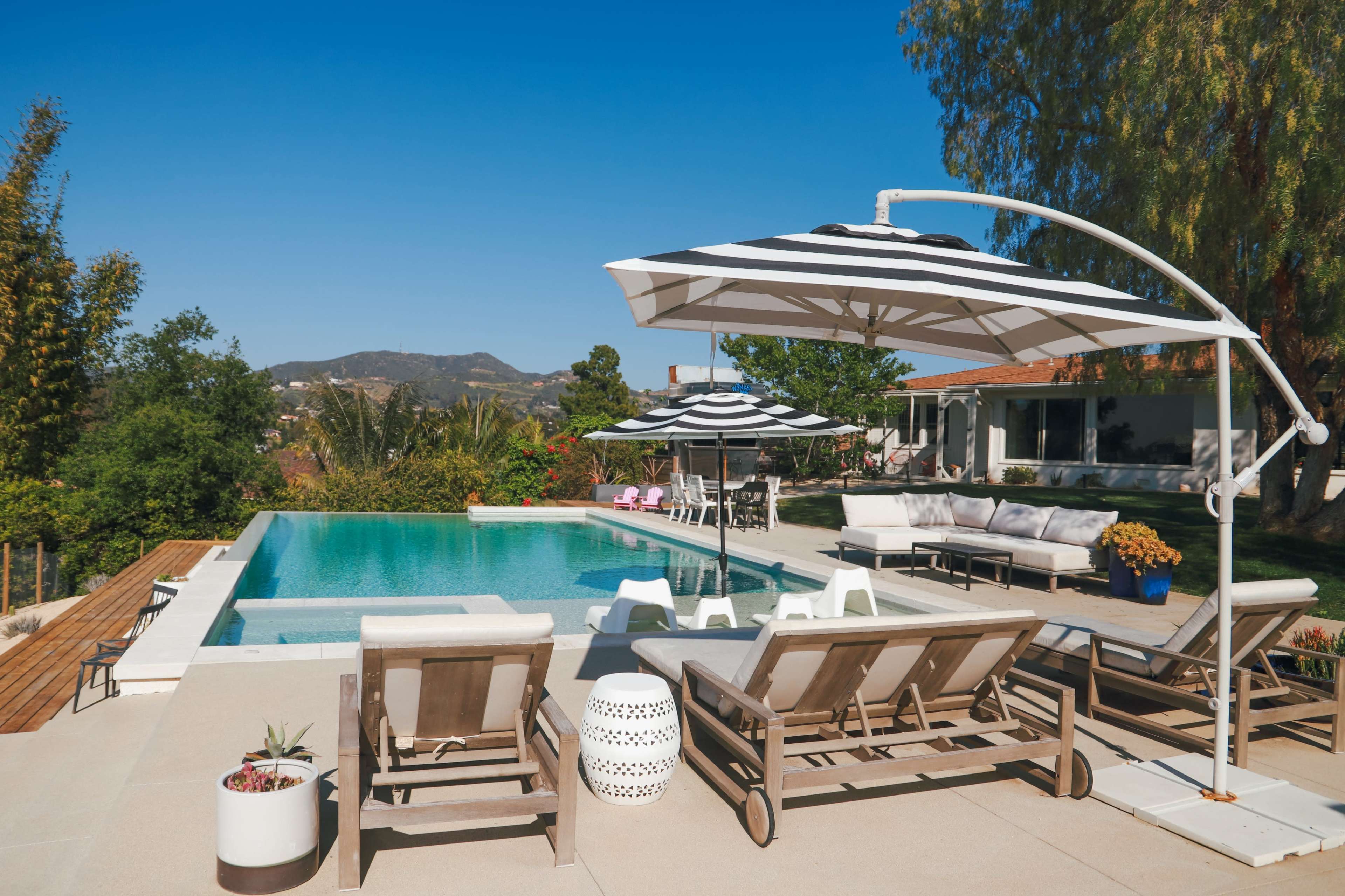 A backyard features a swimming pool surrounded by lounge chairs and large striped umbrellas, with a view of hills in the background.