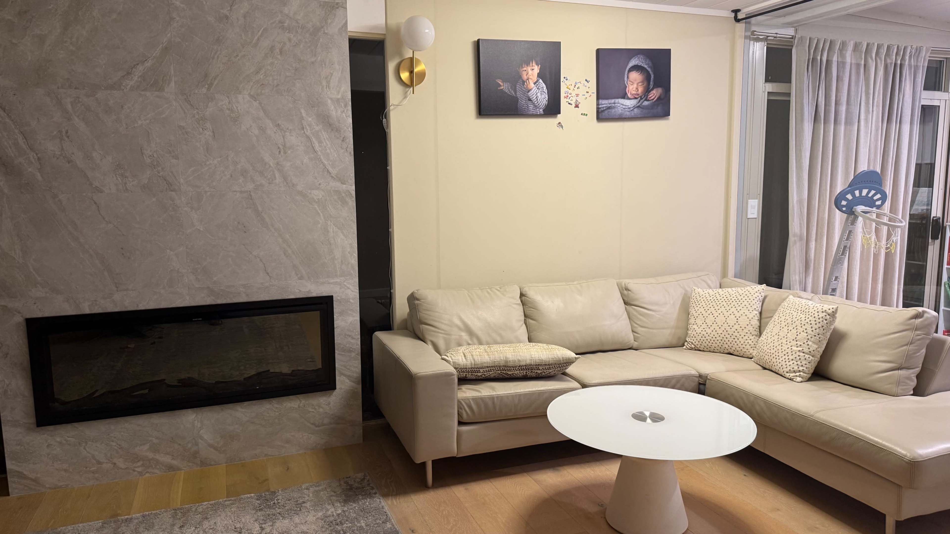 The image shows a contemporary living room featuring a light-colored sectional sofa, a round coffee table, and a wall-mounted fireplace, along with two framed pictures hung above the sofa.