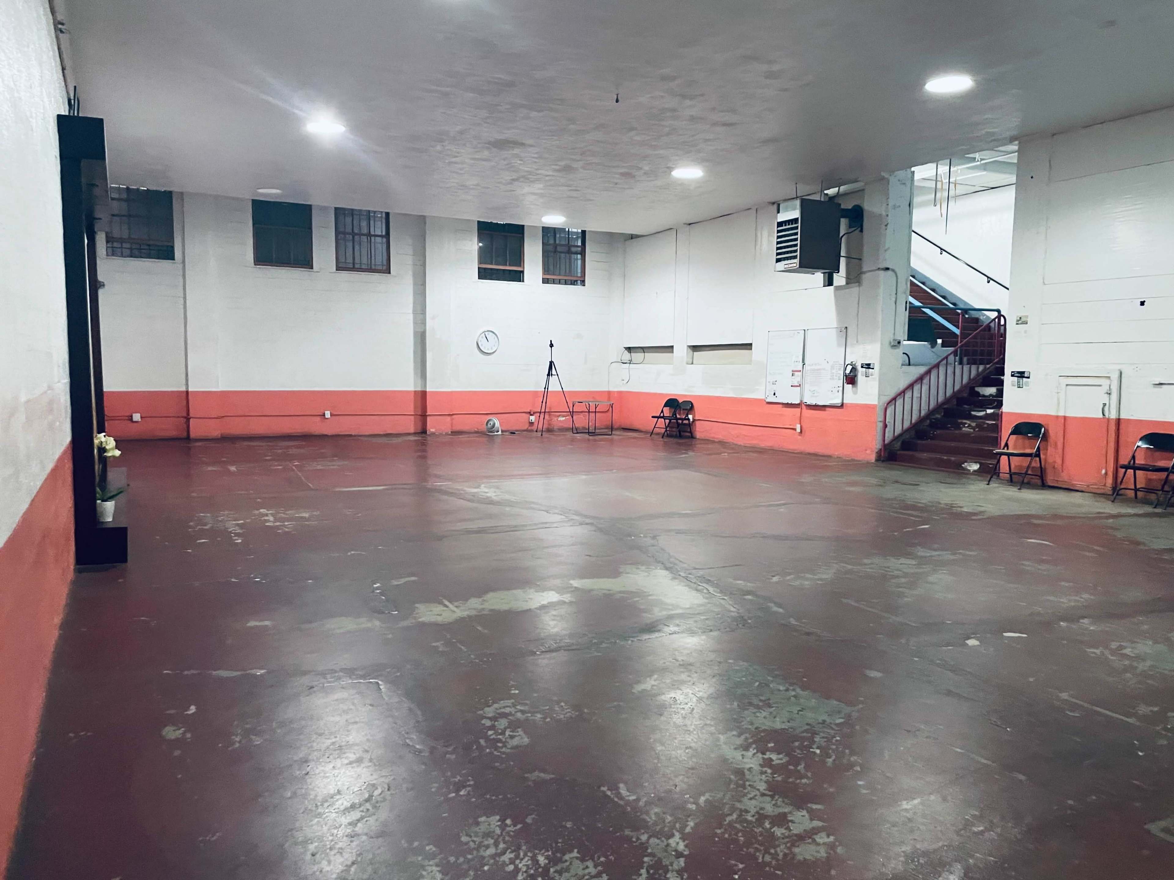 The image shows a spacious, empty room with a polished concrete floor, white walls, and a red stripe along the bottom, featuring a staircase and several chairs positioned against one wall.