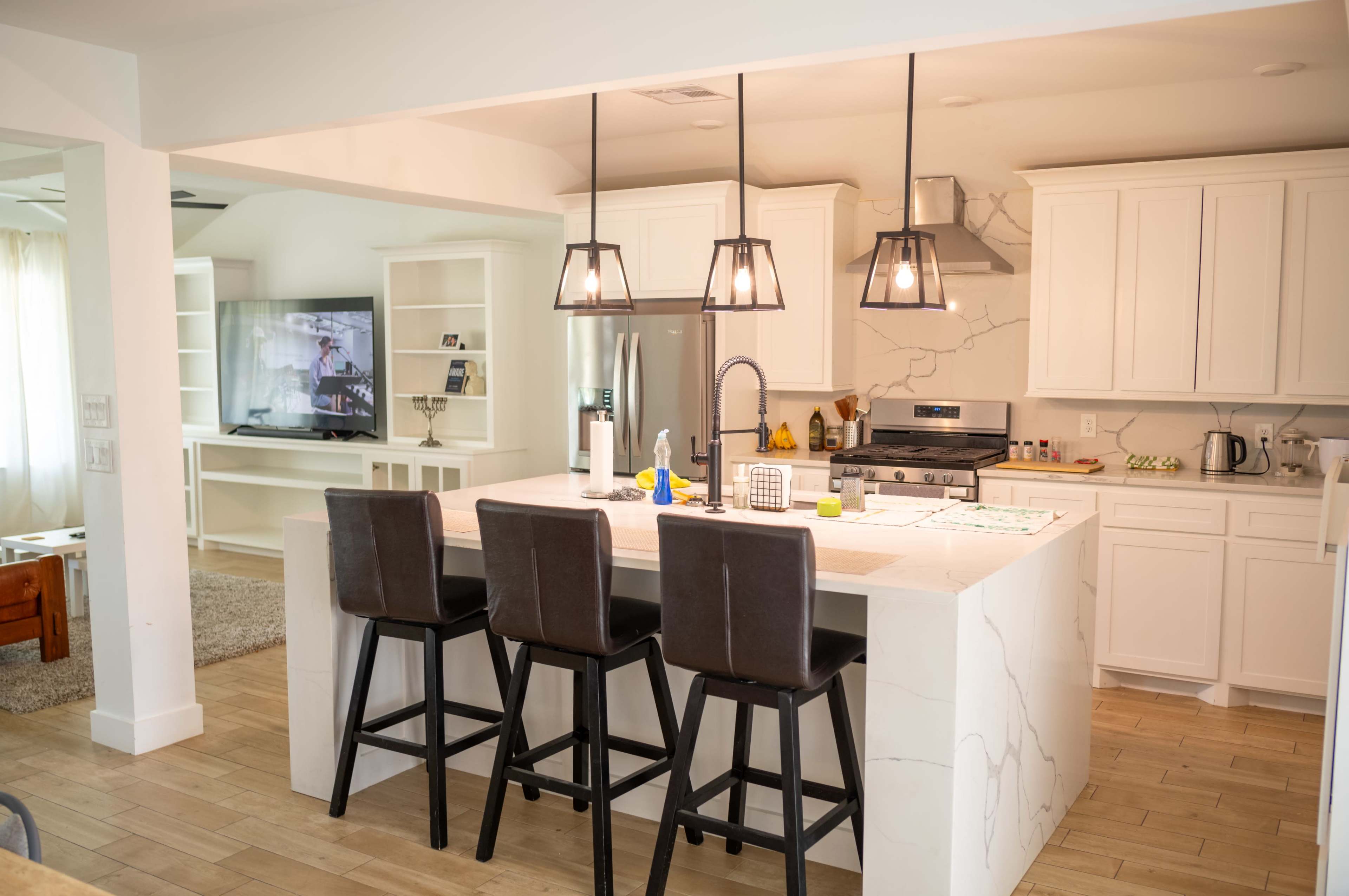 The image shows a modern kitchen with white cabinets, a large island with seating for three, and a television mounted on the wall in the background.