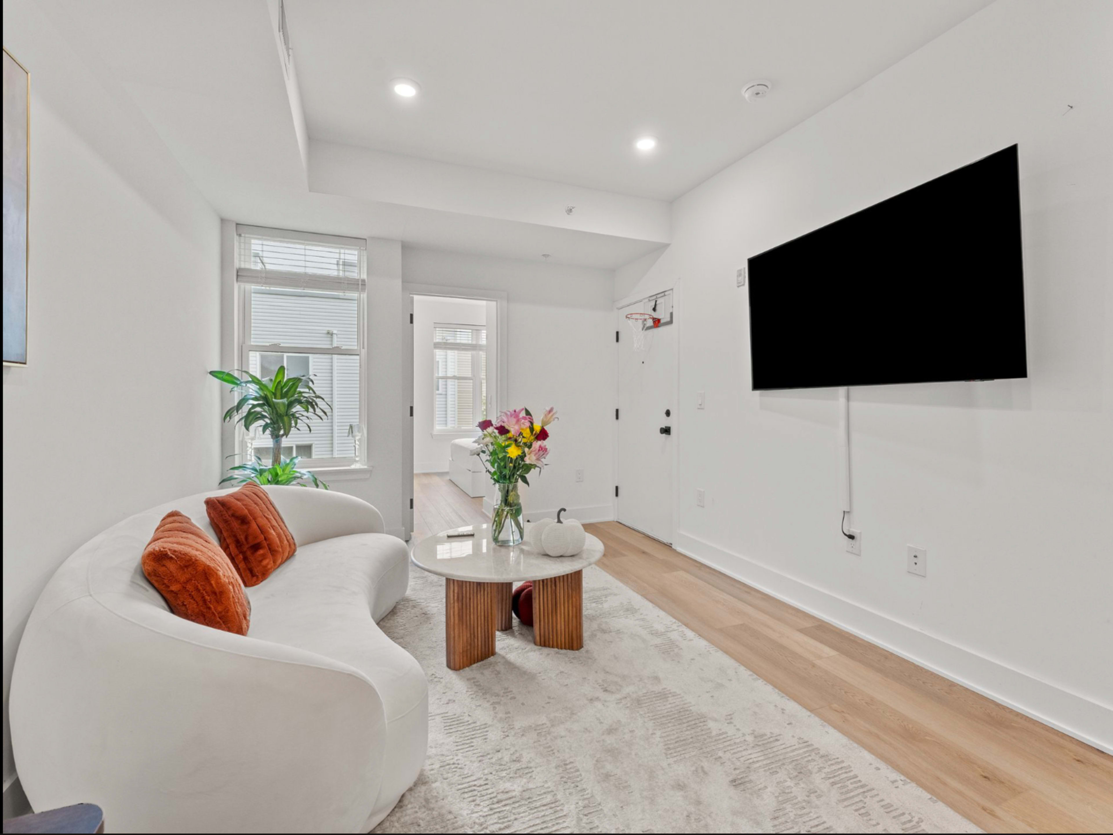A modern living room features a white curved sofa, a wooden coffee table with a vase of flowers, and a wall-mounted television.