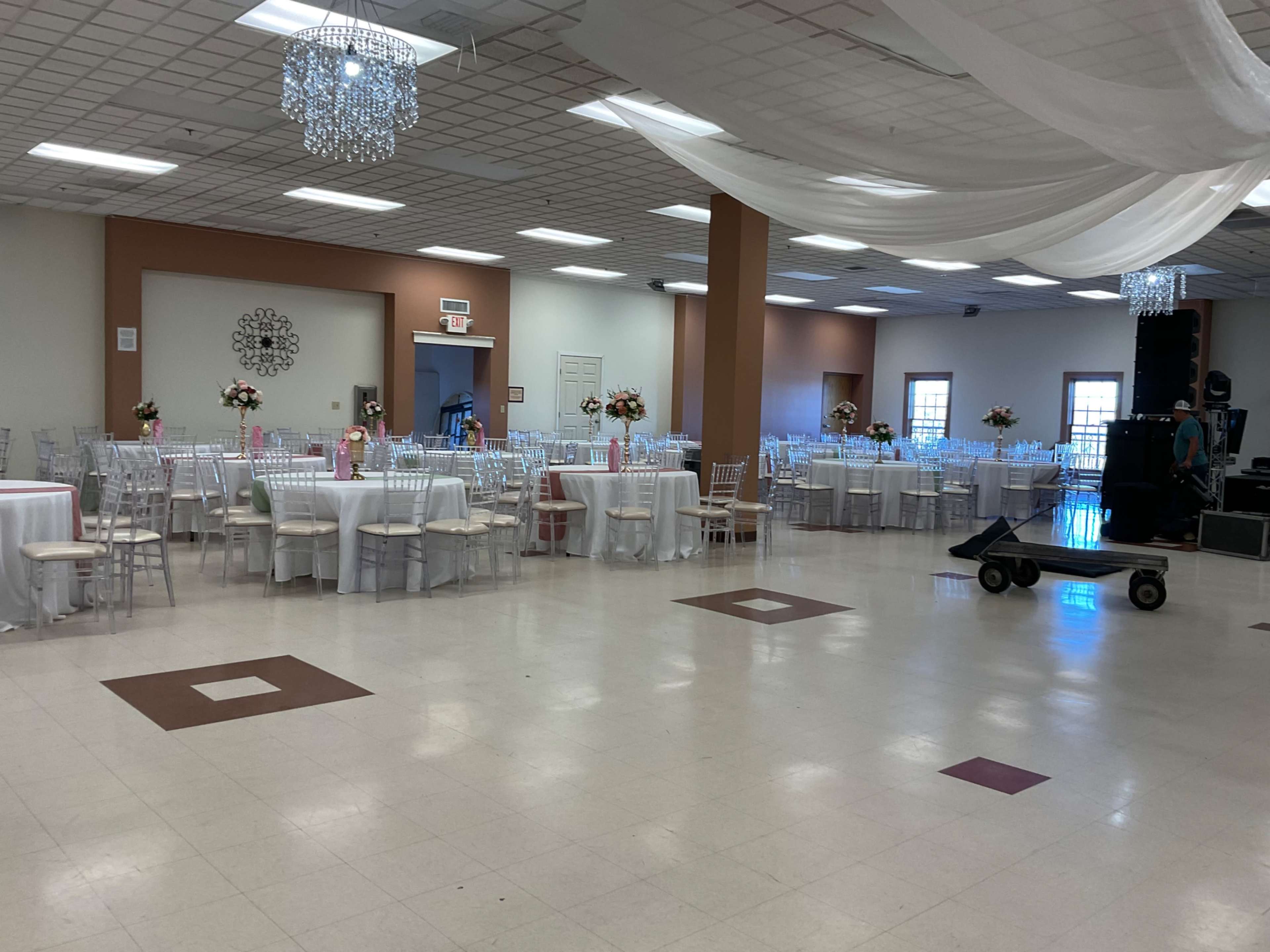 The image shows a wide view of a banquet hall set up for an event, with neatly arranged tables and chairs, floral centerpieces, and a dance floor area.