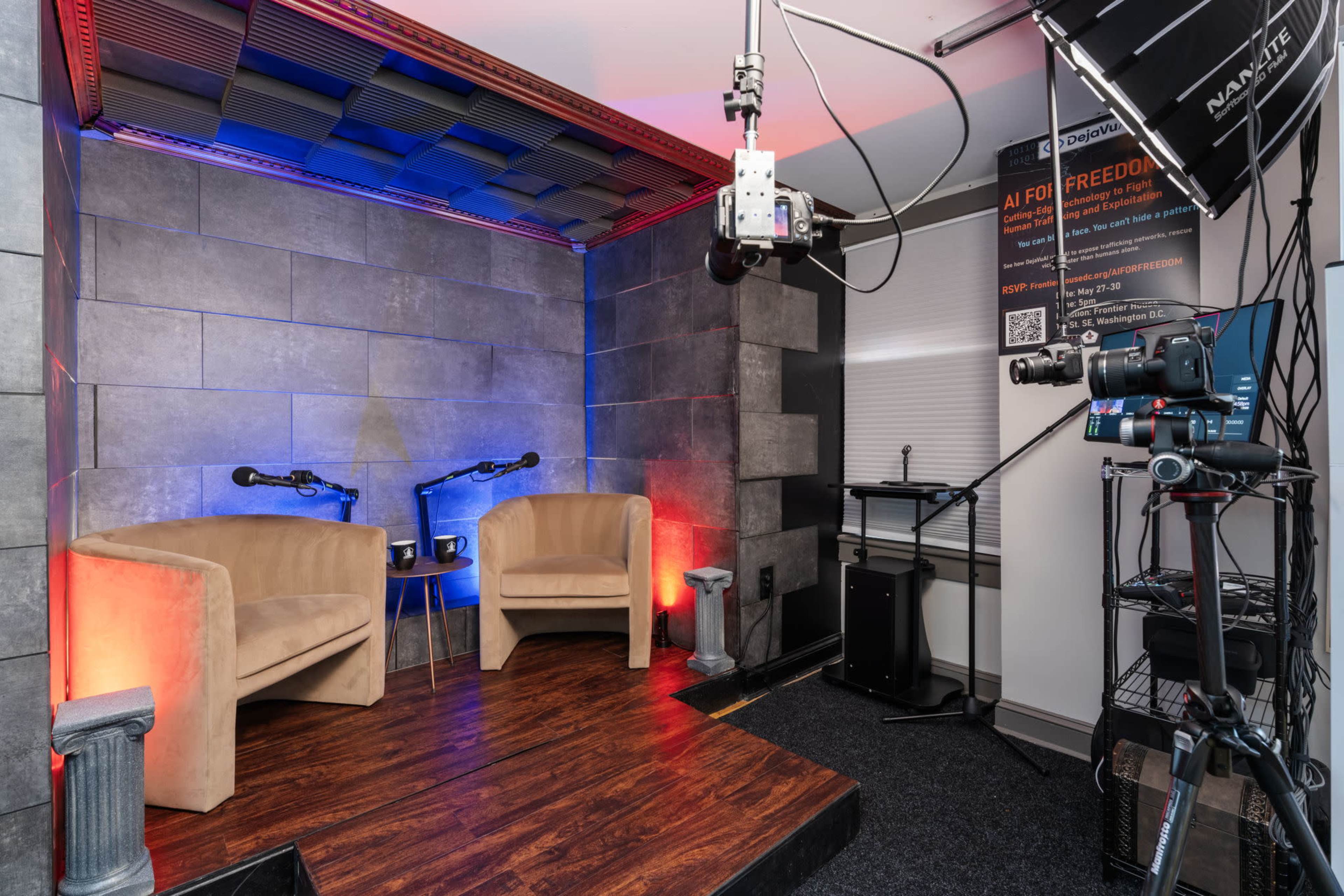 The image shows a video studio set up with two chairs, microphones, and camera equipment against a backdrop of textured gray walls and colored lighting.