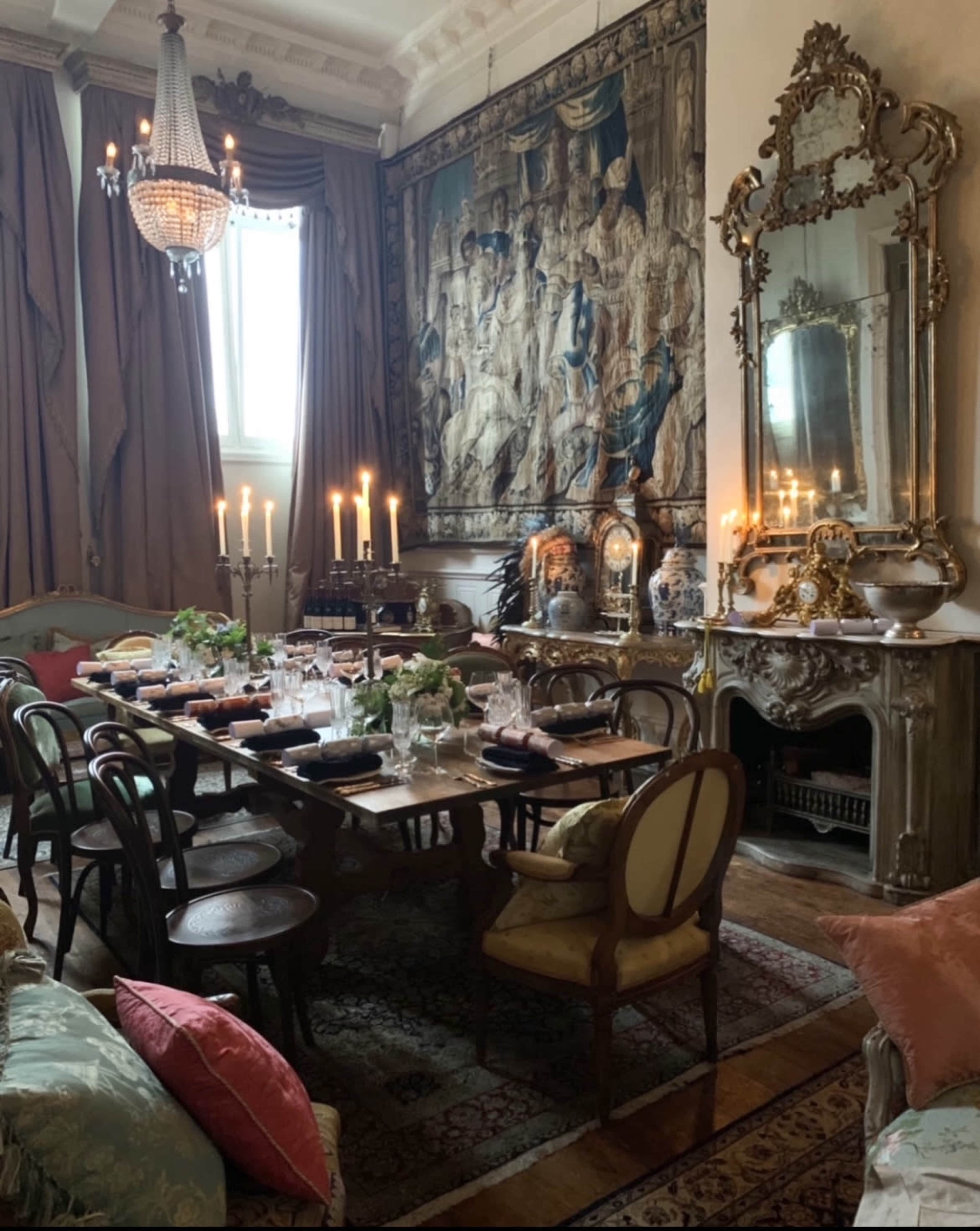 The picture shows an elegantly arranged dining room featuring a large wooden table set with dishes and candles, surrounded by various seating and decorated with a tapestry and a mirror.