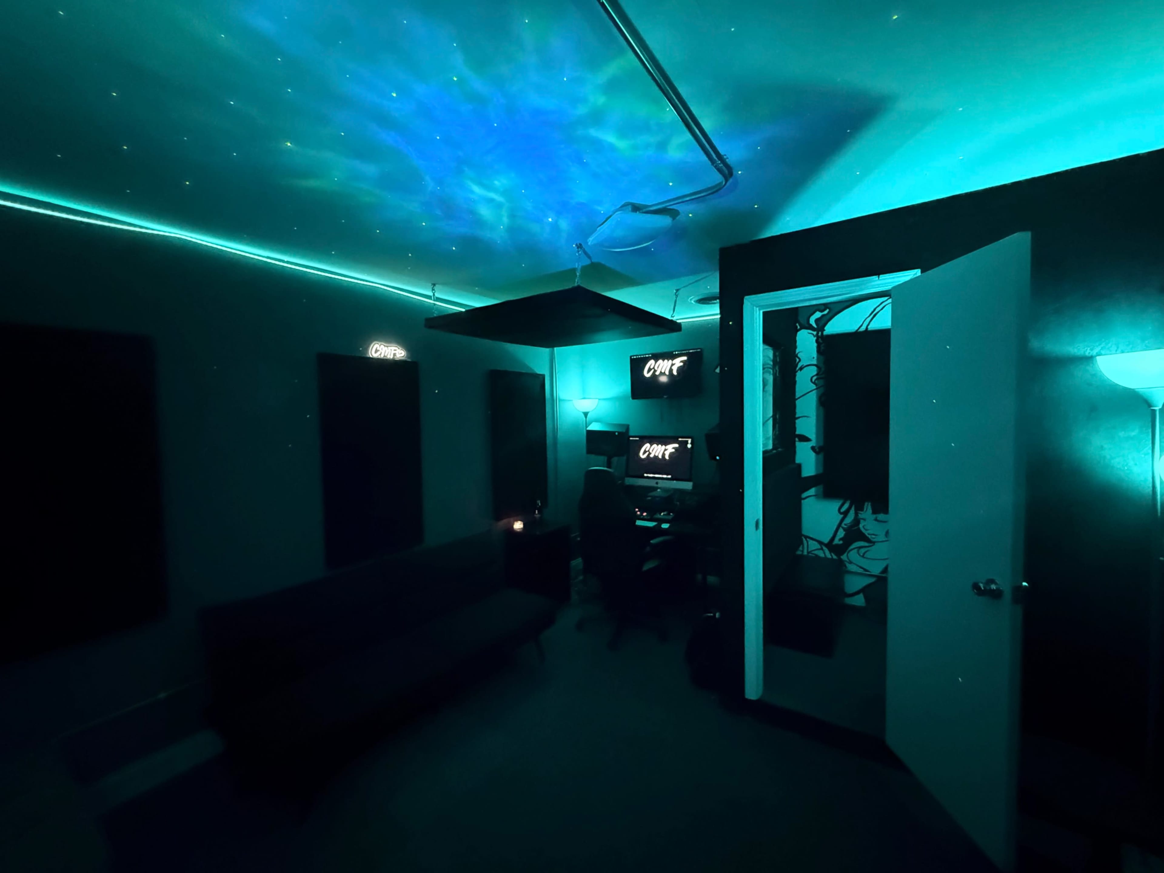The image shows a dimly lit room with teal lighting, featuring a couch, two computer screens, and star-like projections on the ceiling.