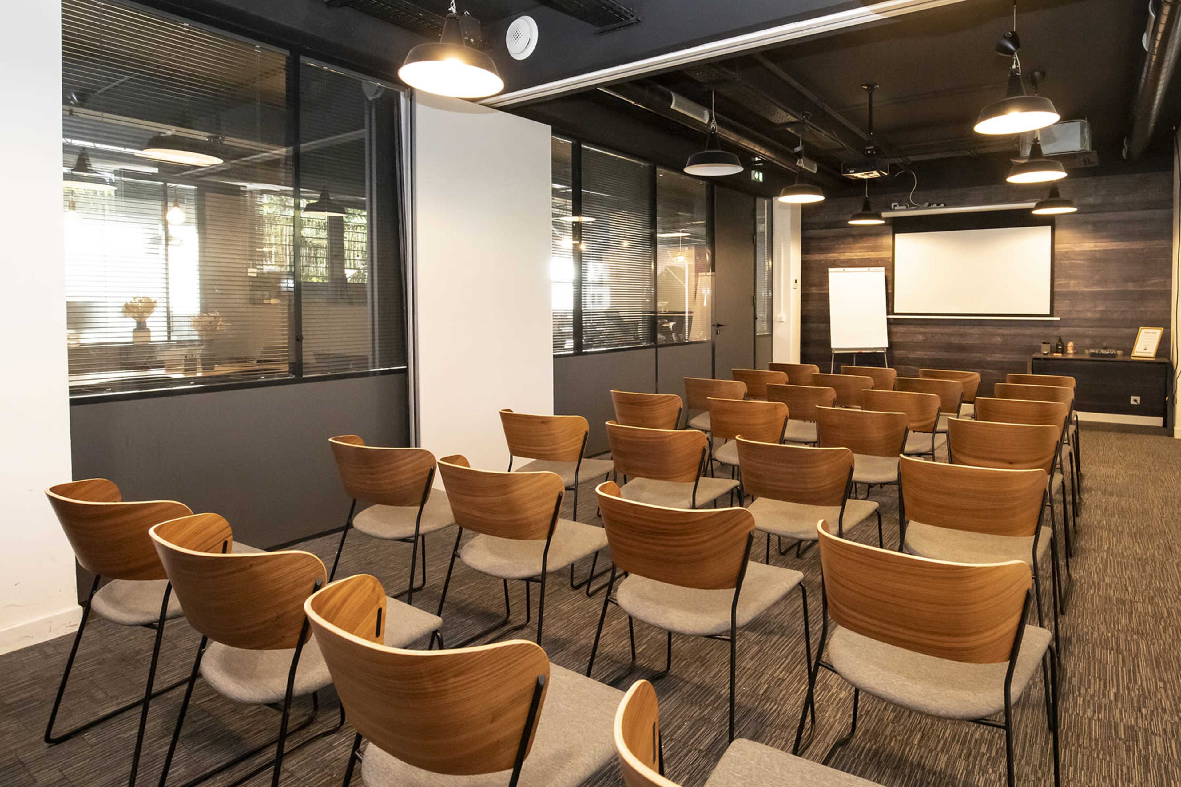 A conference room set up with rows of wooden and fabric chairs facing a projection screen, with large windows and modern lighting fixtures.