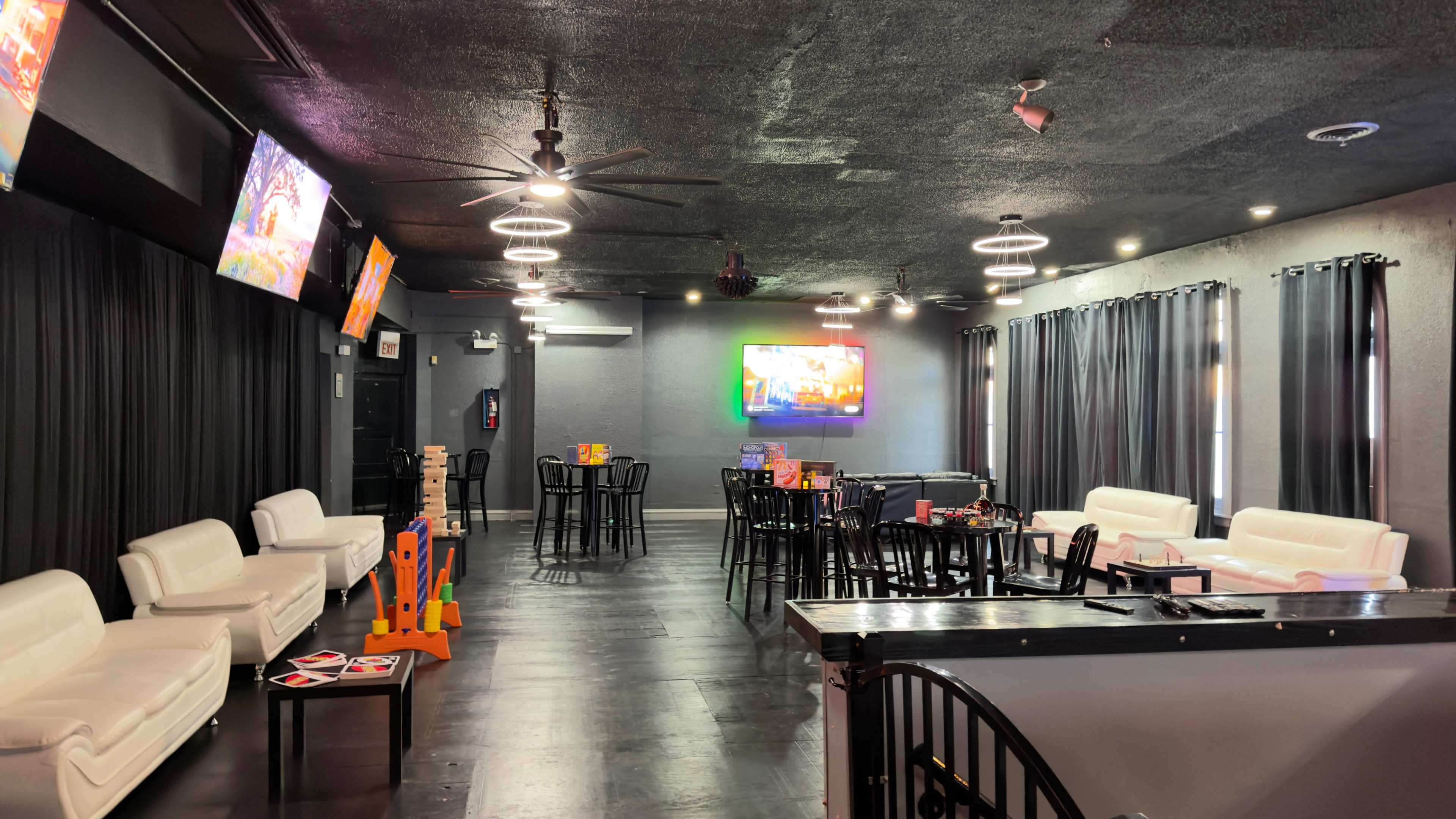 The image shows a modern lounge area with white sofas, black tables, and several flat-screen TVs on the walls, featuring dim lighting and dark curtains.