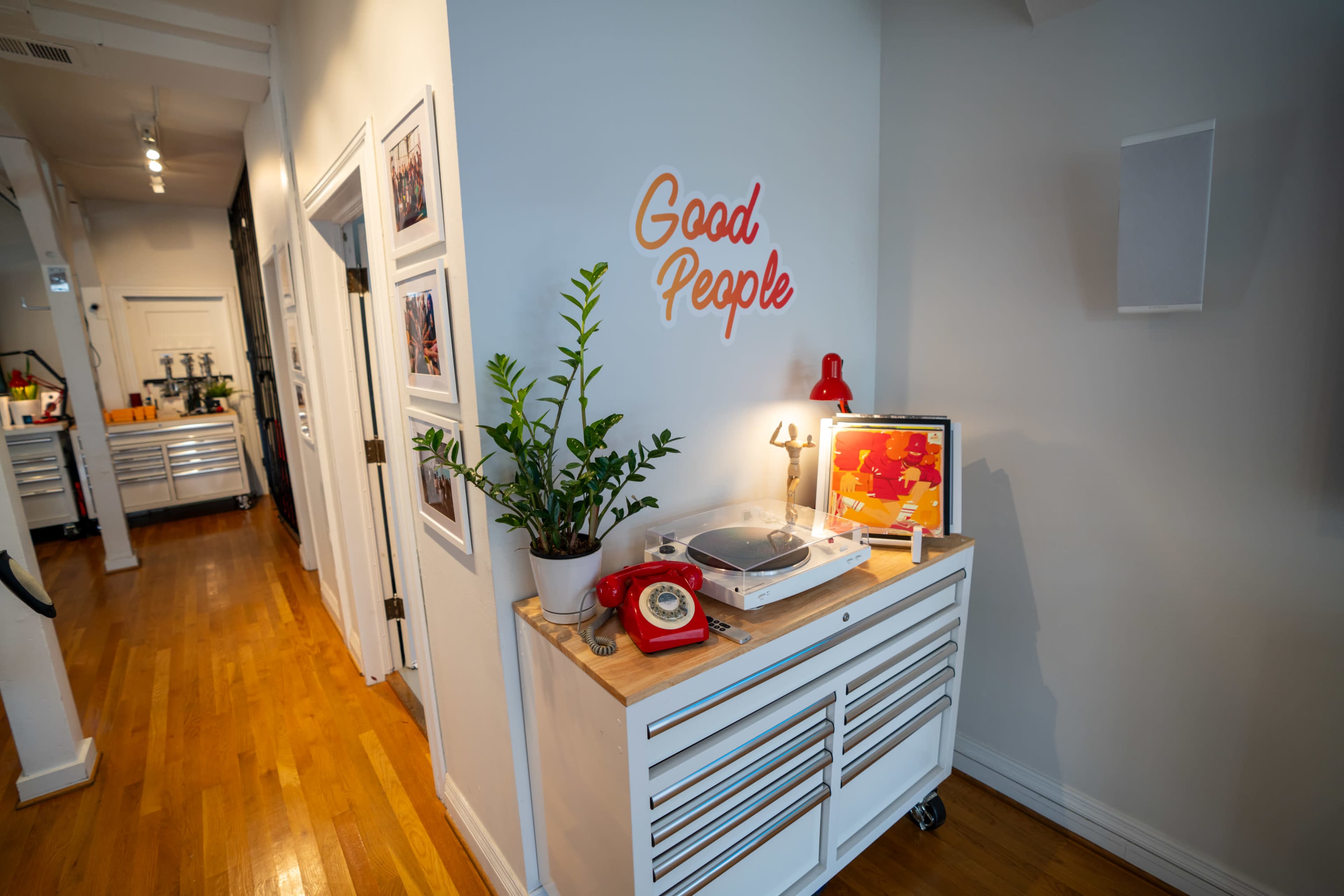 A narrow hallway features a console table with a record player, a potted plant, and a vintage rotary phone, alongside a bright red sign that reads "Good People."