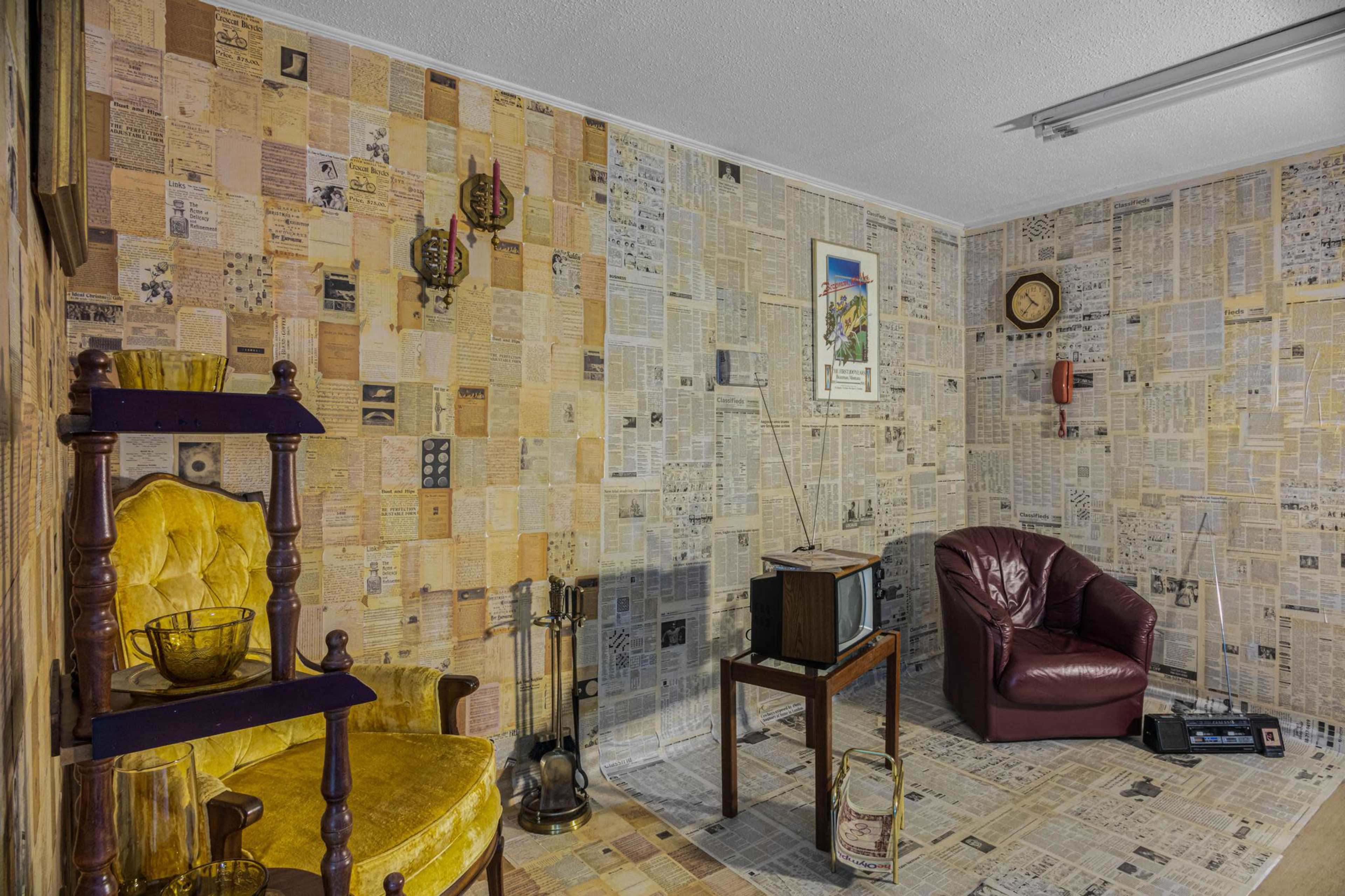 The room features walls covered in newspaper clippings, with a yellow armchair, a maroon leather chair, a wooden table, and a vintage television set.