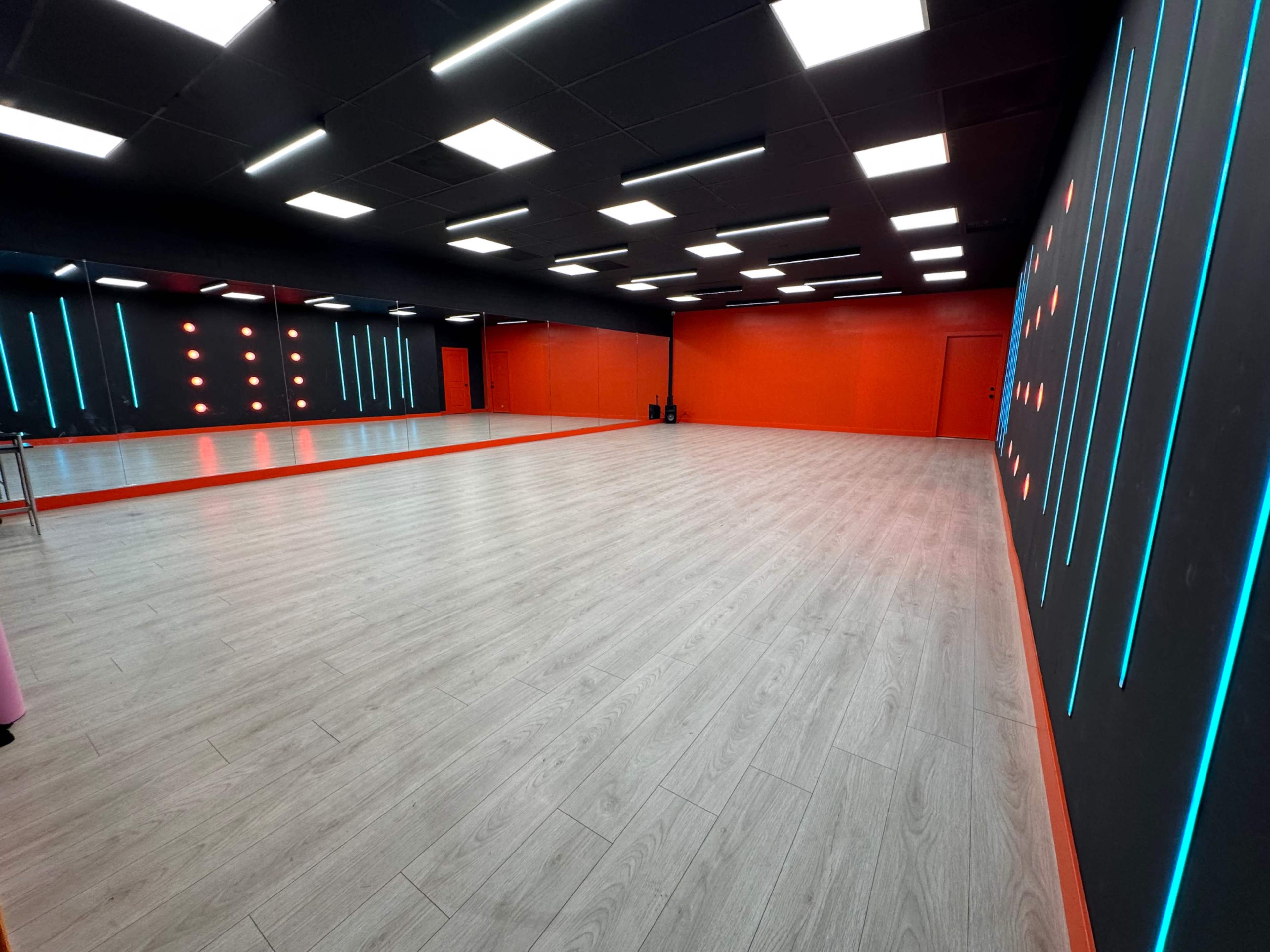 A spacious dance studio featuring a wooden floor, orange walls, and illuminated blue and red accents.