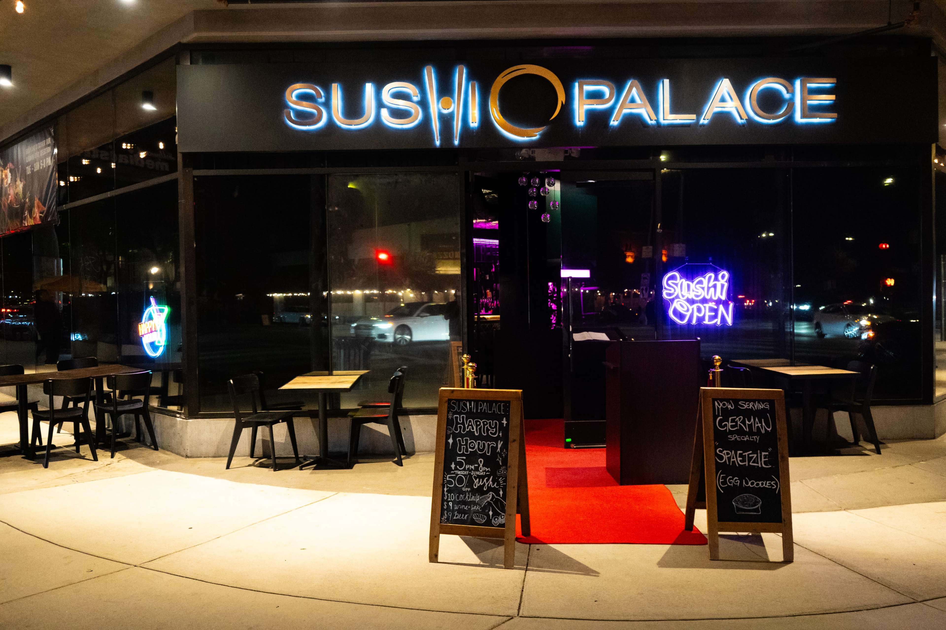 The image shows the entrance of a sushi restaurant named "Sushi Palace," featuring a red carpet, outdoor seating, and illuminated signs indicating it is open.