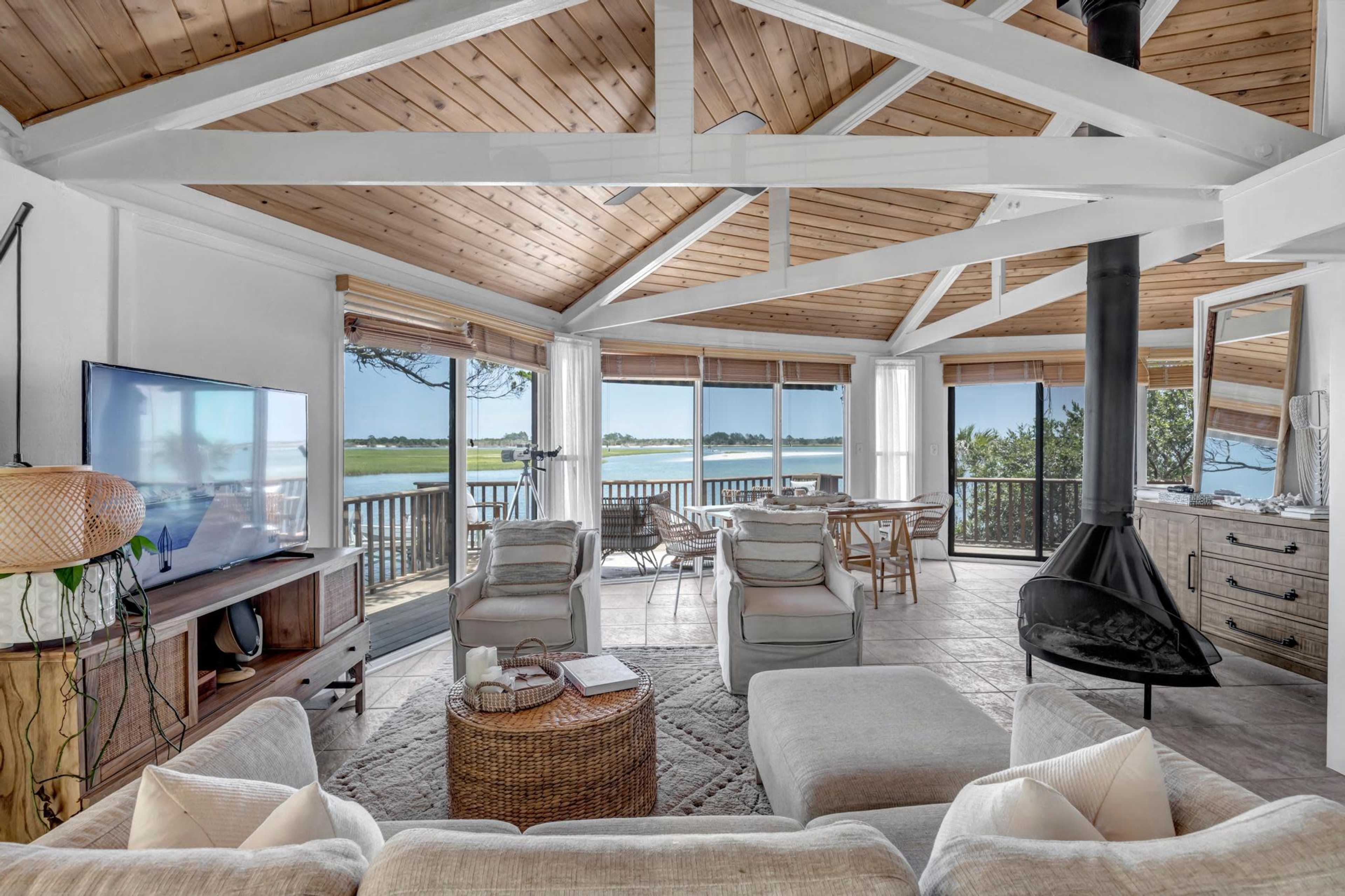 A spacious living room features a large window with a view of the water, complemented by wooden beams and a modern fireplace.