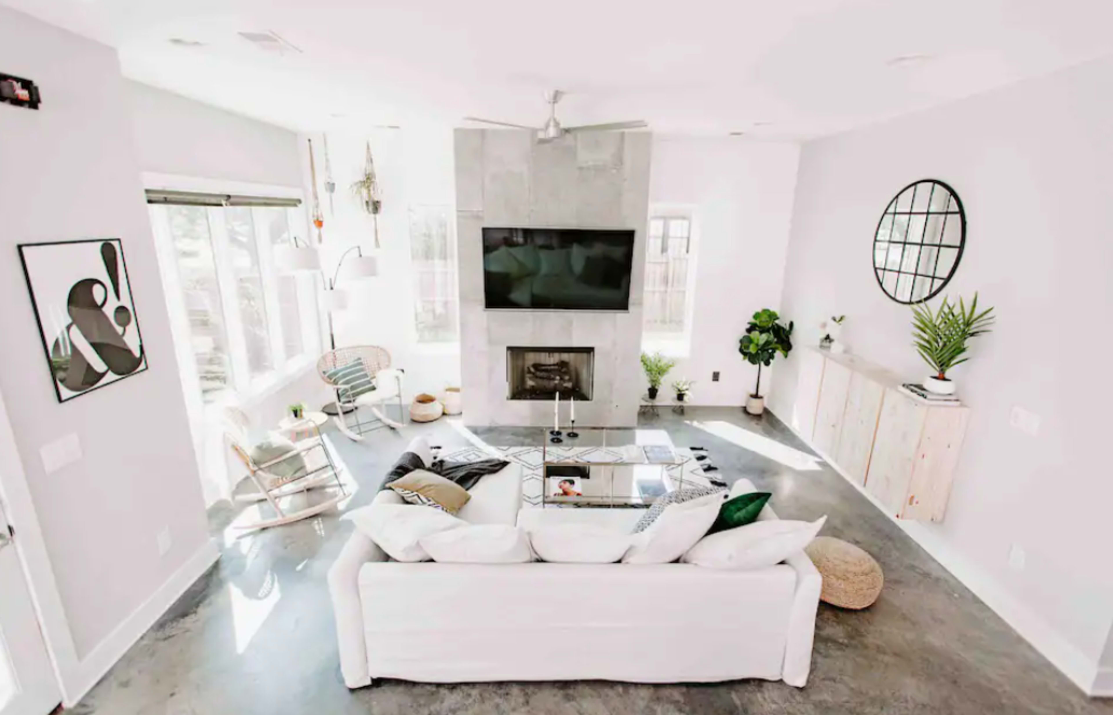 The image shows a modern living room featuring a white sectional sofa, a flat-screen TV mounted above a stone fireplace, and large windows that allow natural light to fill the space.