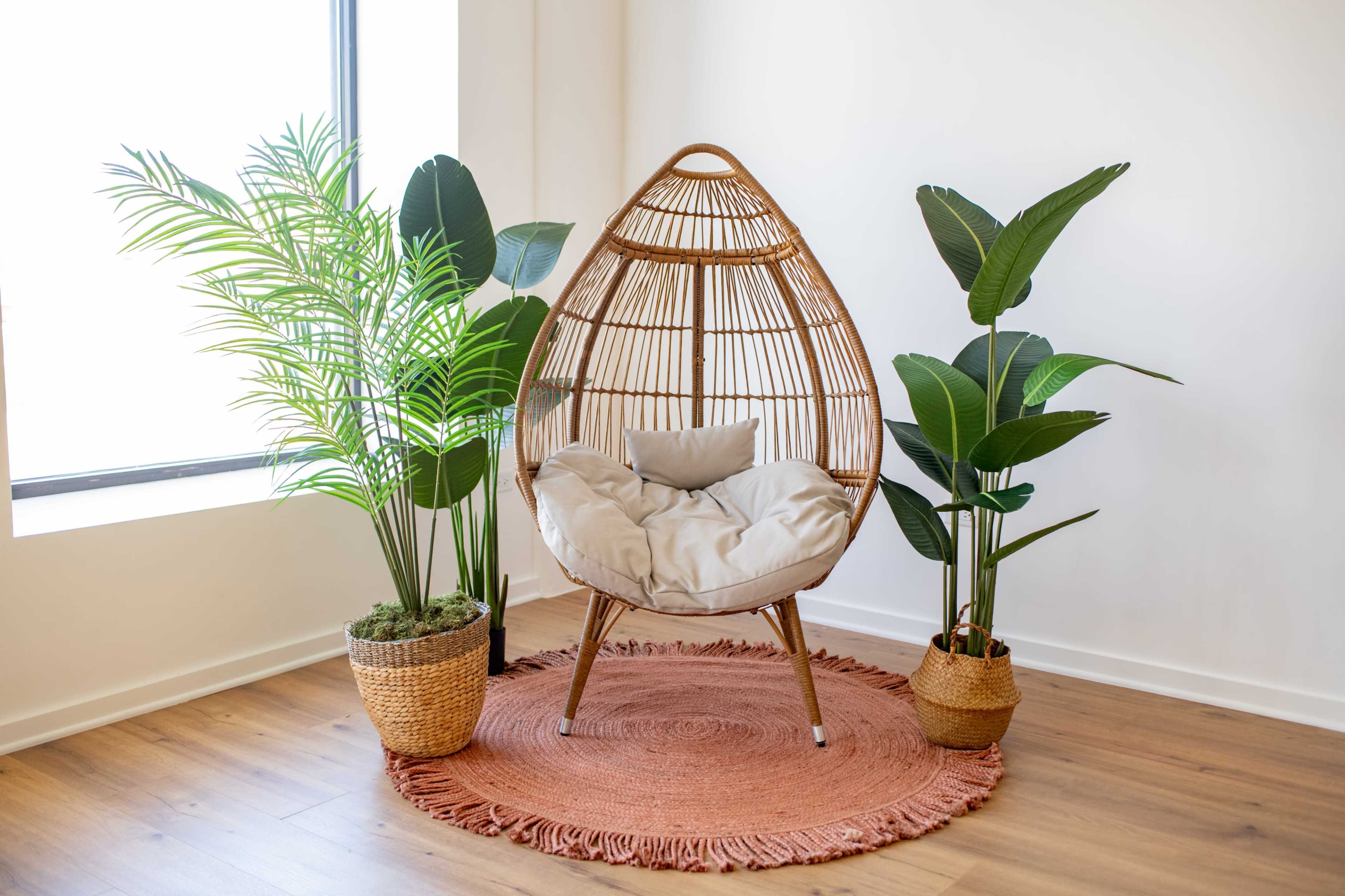 A woven hanging chair with a cushion is placed next to two potted plants on a circular rug in a well-lit room.
