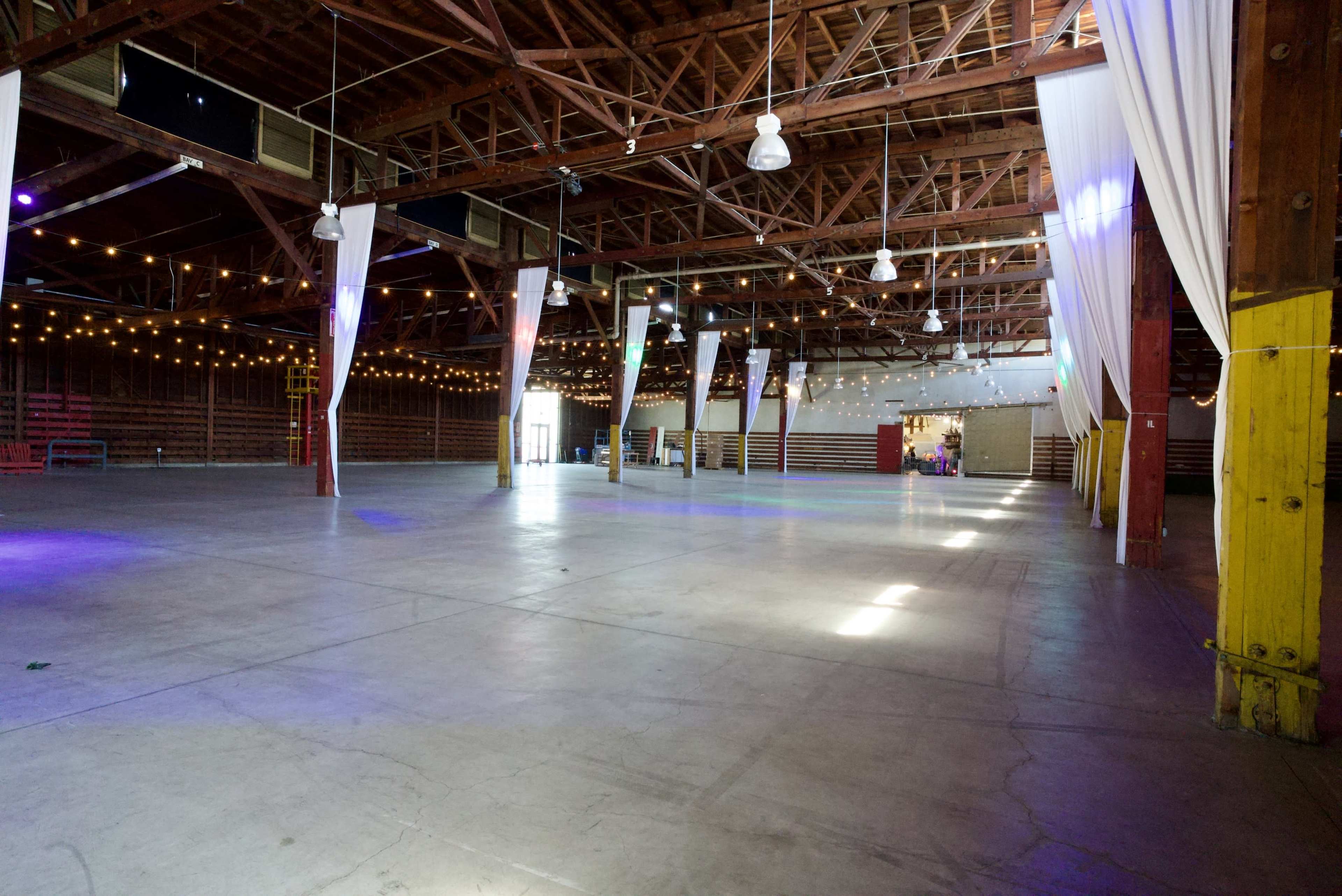 A spacious, vacant warehouse with wooden beams, string lights, and banners hanging from the ceiling.