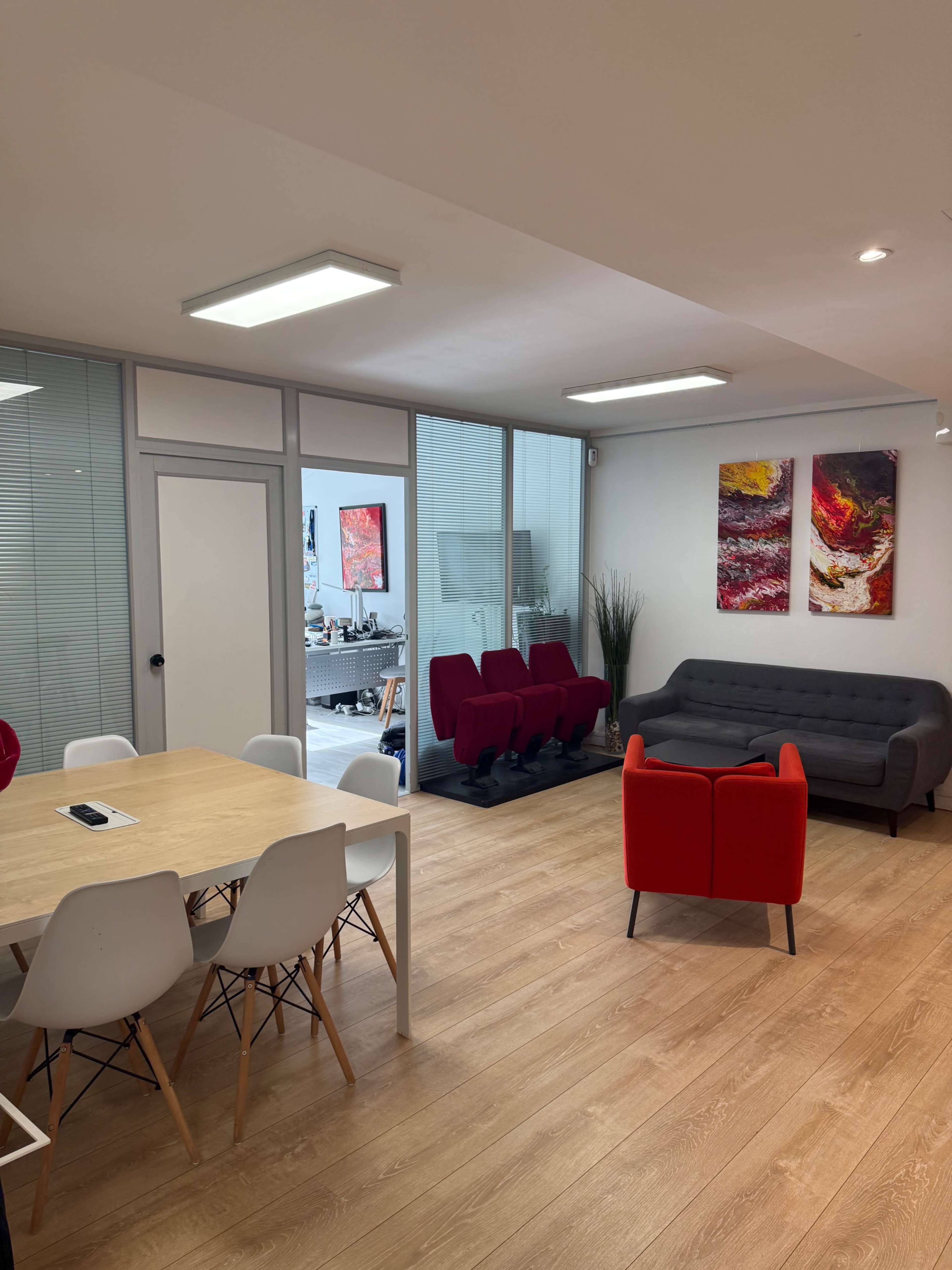 The image shows a modern office space featuring a wooden dining table, a gray sofa, red armchairs, and two abstract art pieces on the walls.