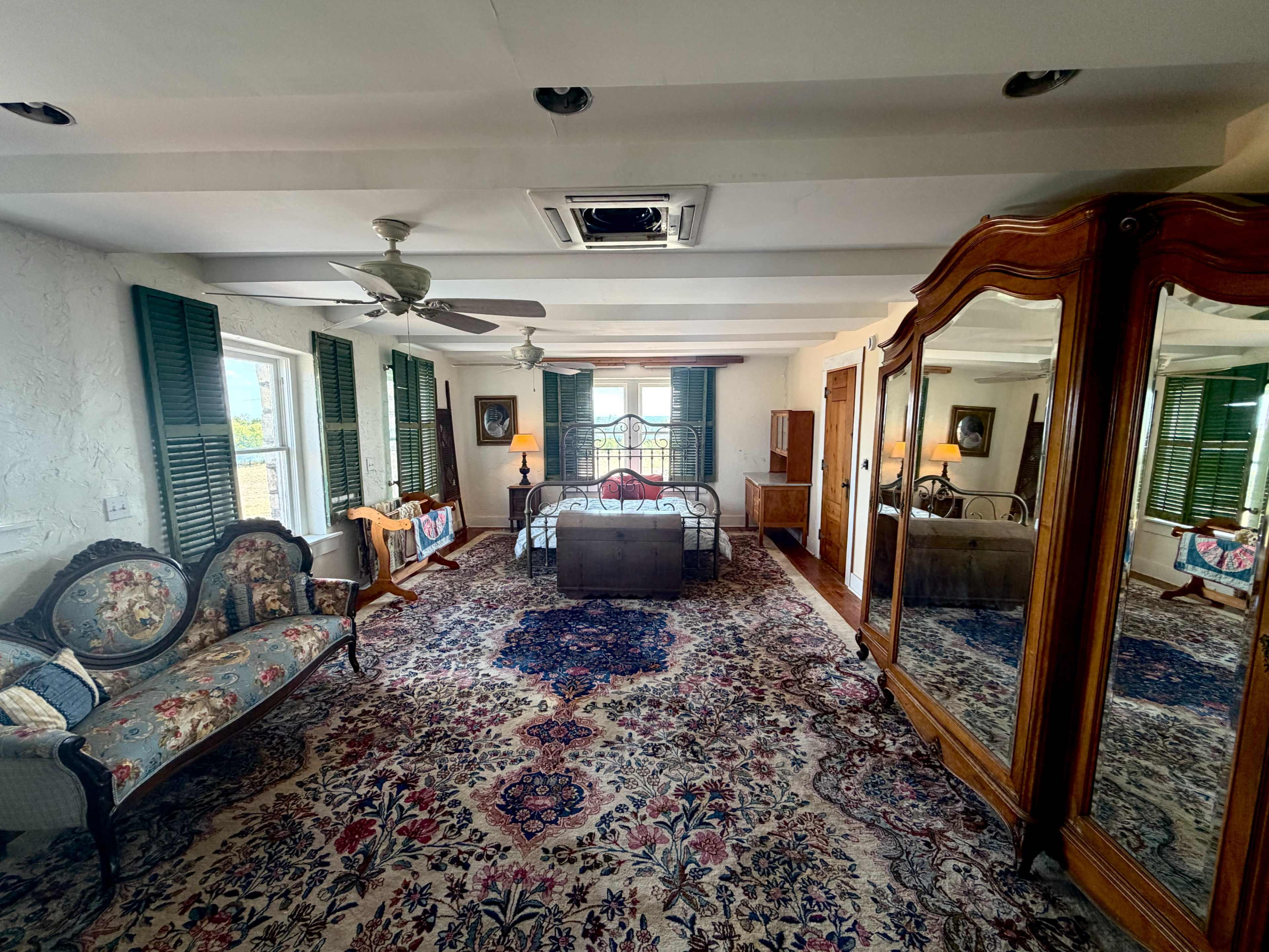 The room features a bed, a vintage seating area, dark green window shutters, and an ornate mirror wardrobe, all set on a patterned rug.