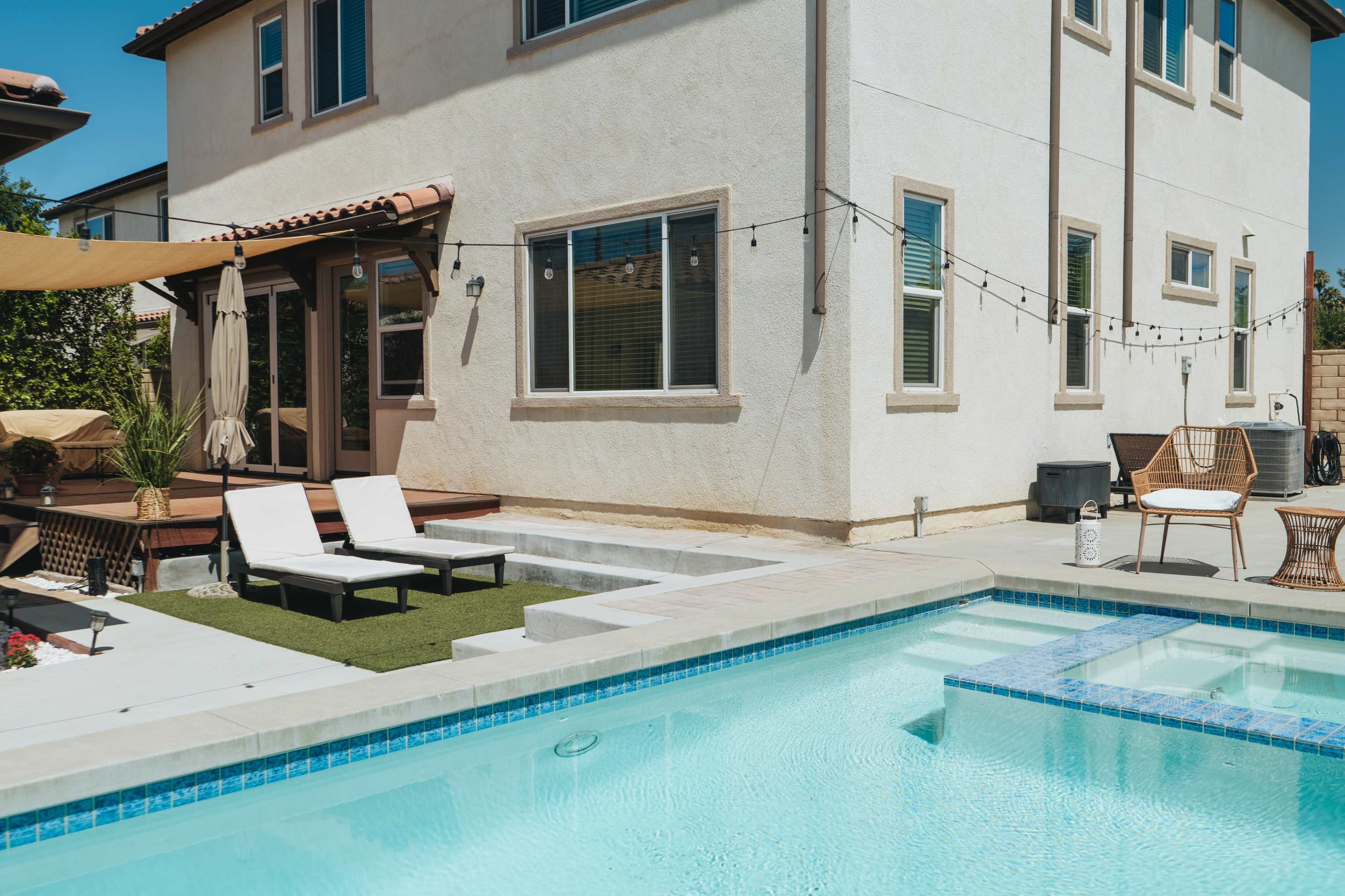 A backyard with a swimming pool, lounge chairs, and a wooden deck area, featuring string lights and a patio umbrella.
