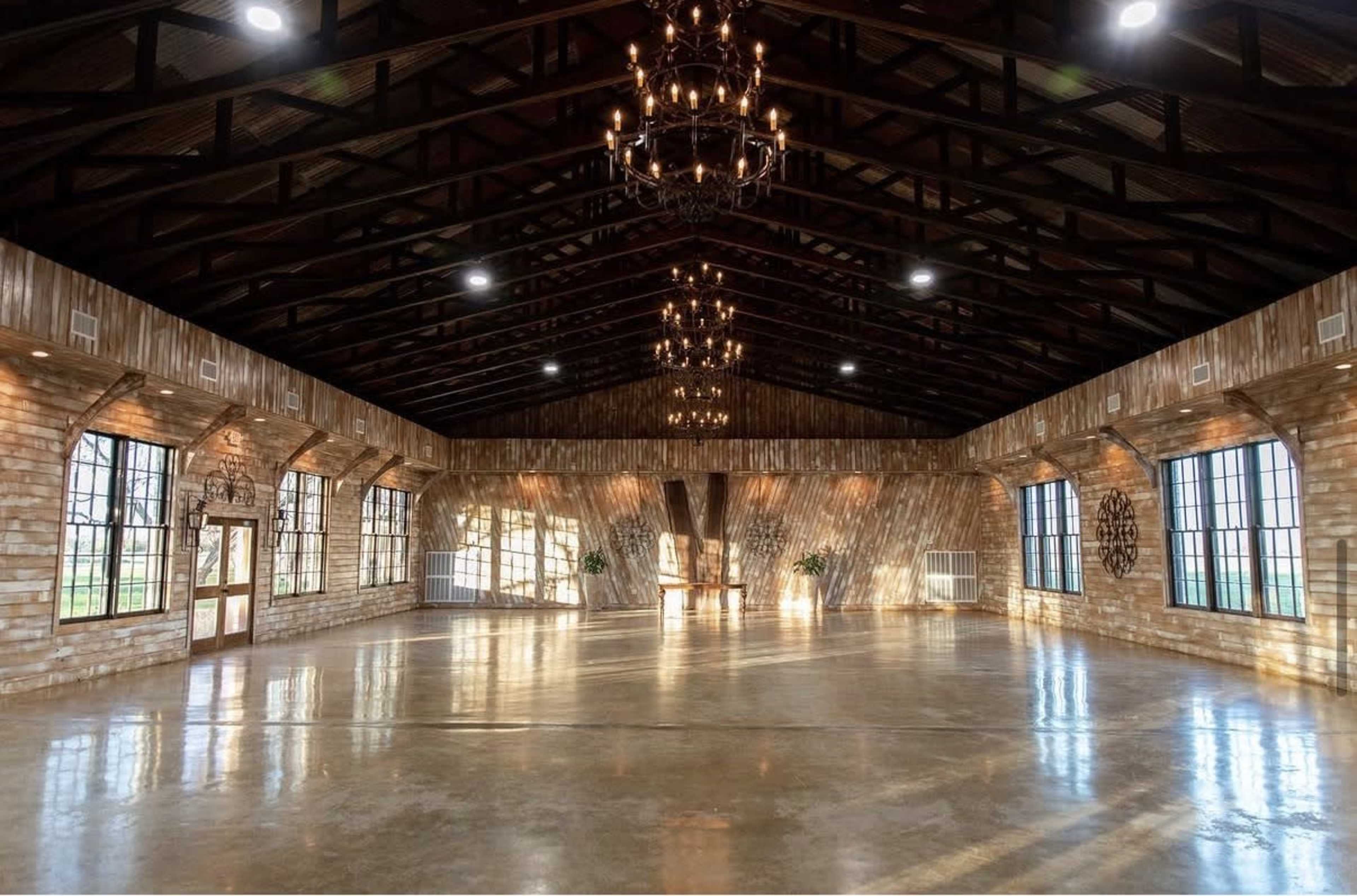 The image shows a spacious indoor venue with high ceilings, large windows, and wooden beams, featuring polished concrete floors and stylish chandeliers.