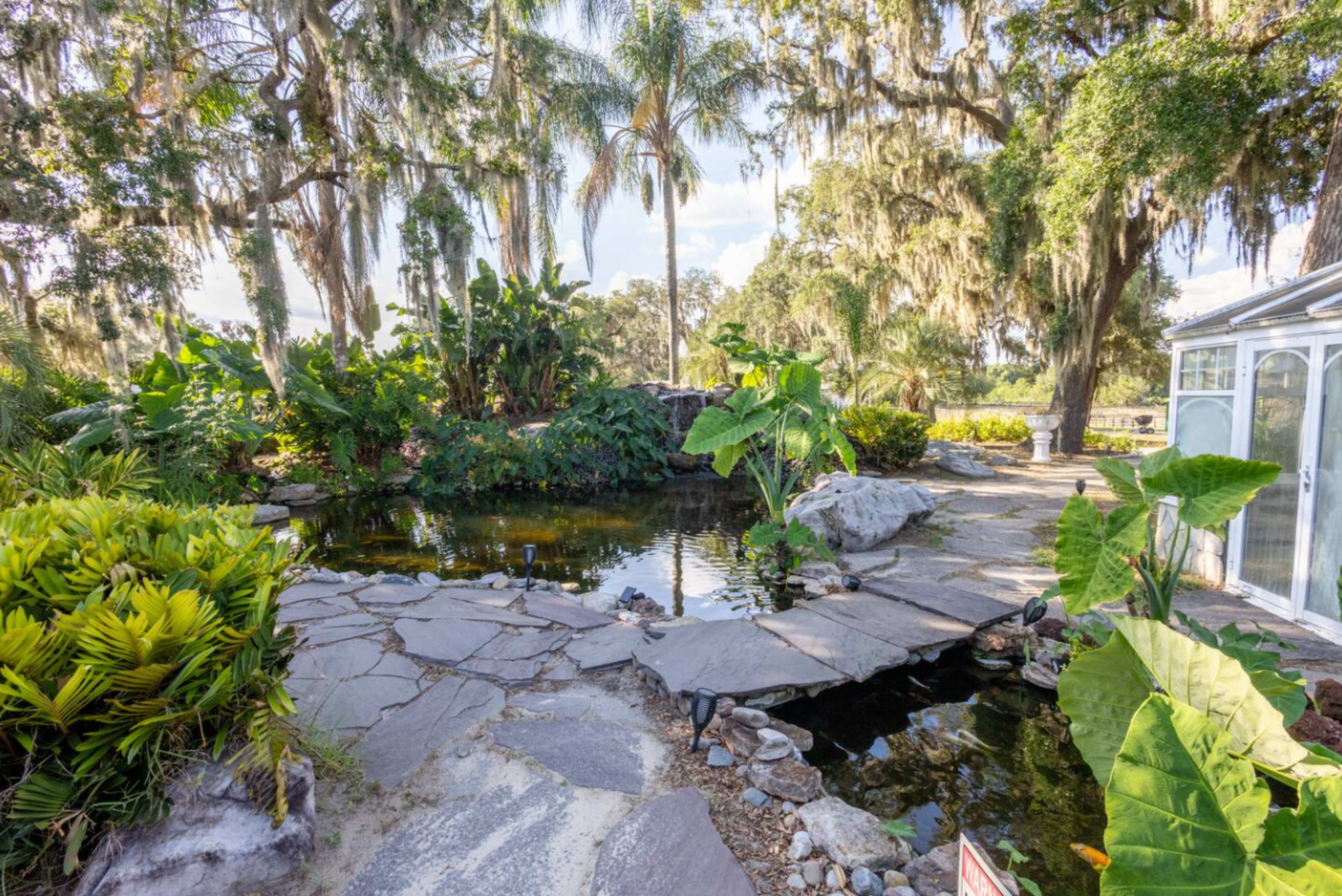 The image shows a landscaped garden with a stone pathway leading over a pond surrounded by lush greenery and palm trees.