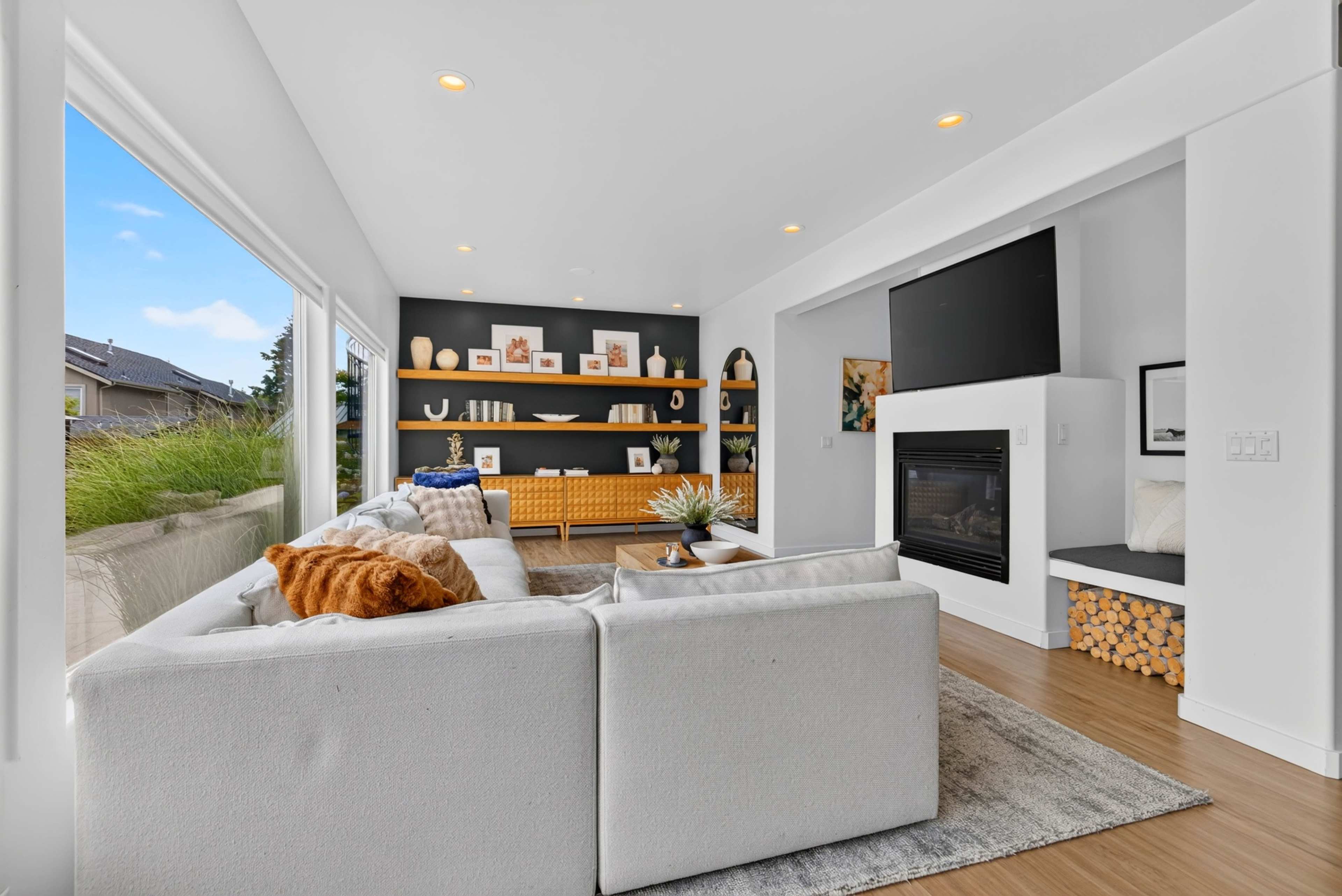 A modern living room features a white sectional sofa, a wall-mounted television, a fireplace, and shelves with decorative items against a dark backdrop, with large windows allowing natural light in.