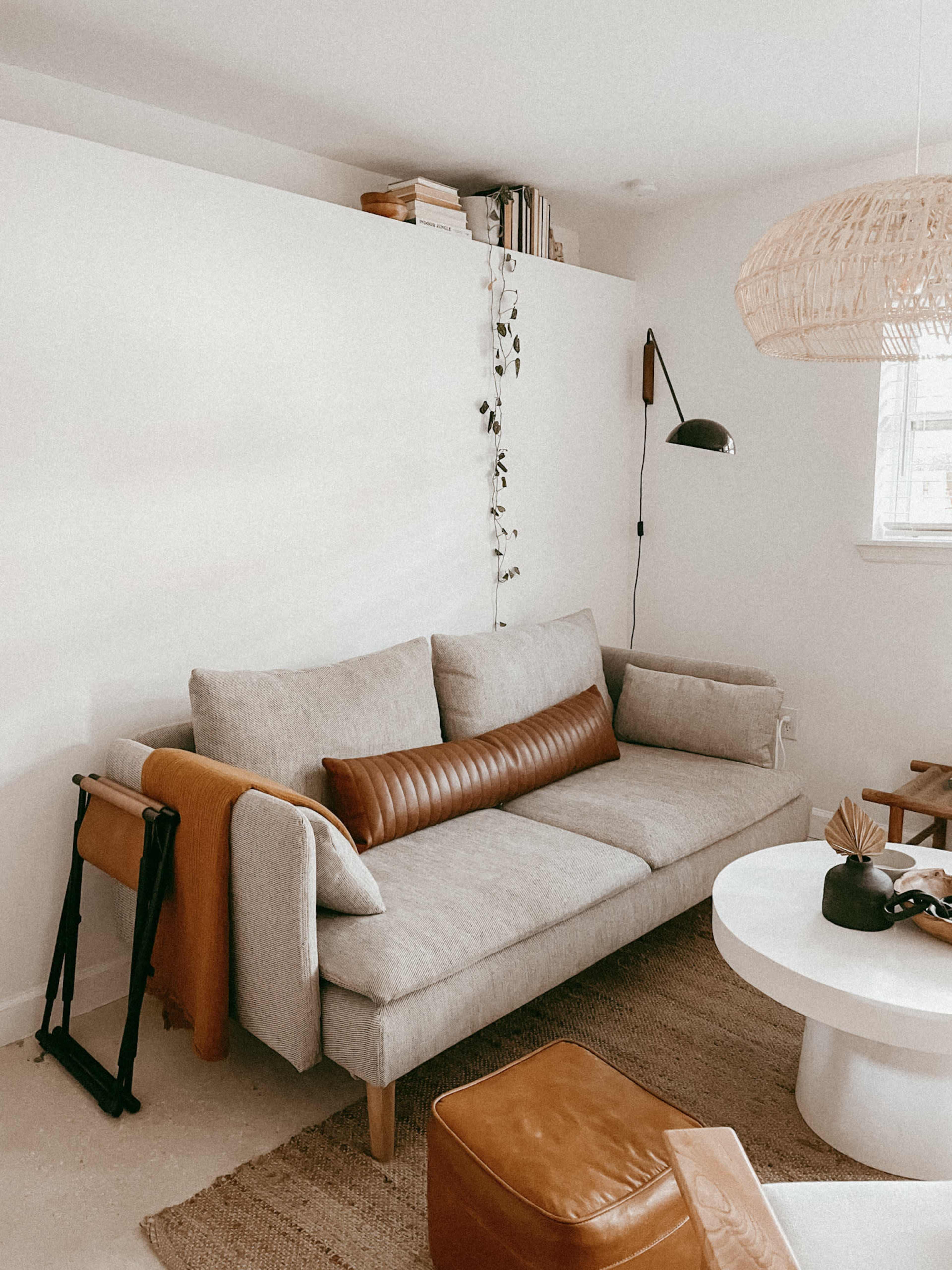 A light-colored living room features a gray sofa with a brown leather pillow, a round white coffee table, and a woven pendant light hanging above.