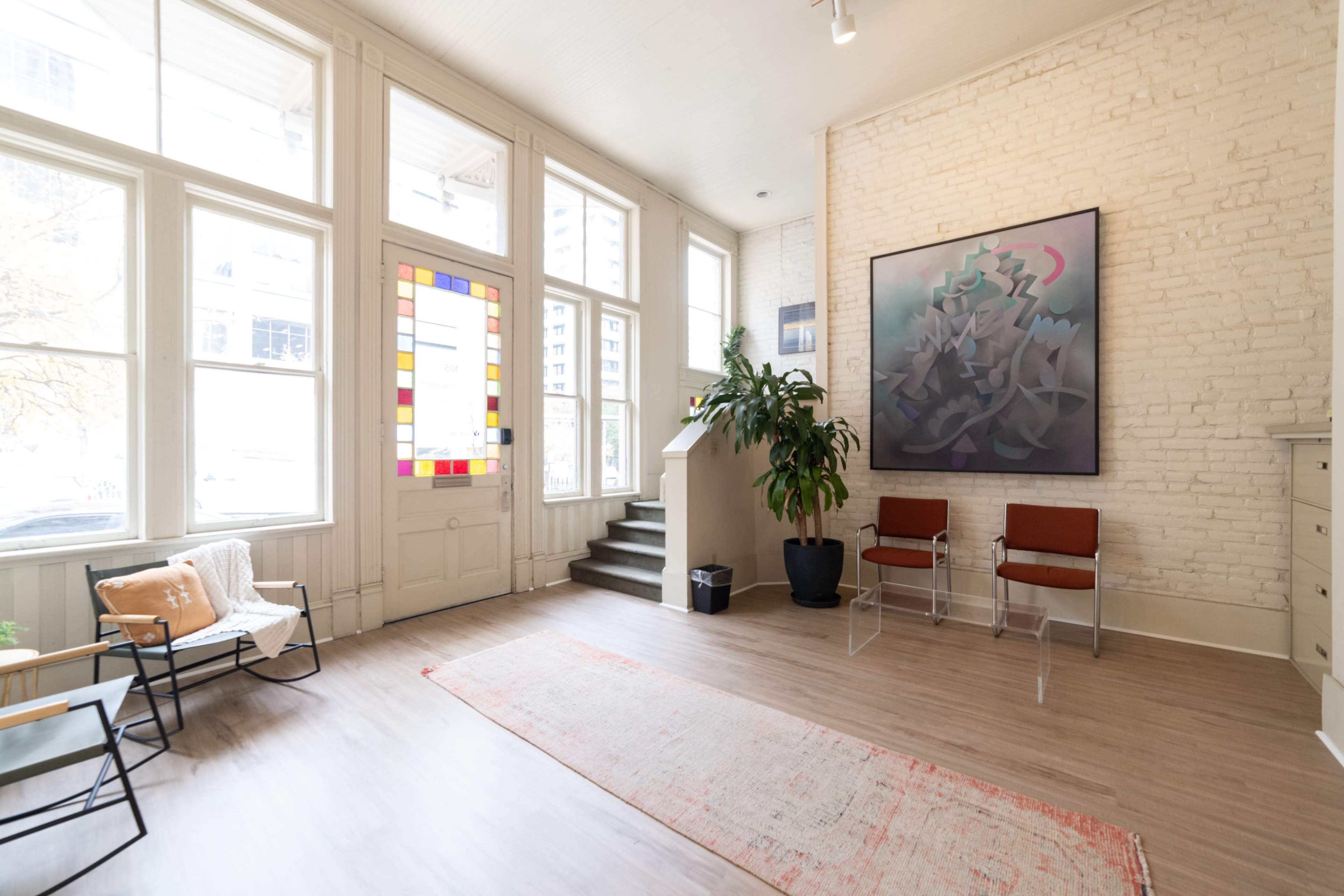 A spacious room with large windows, a colorful stained glass panel, a potted plant, two transparent chairs, and a patterned rug on a wooden floor.