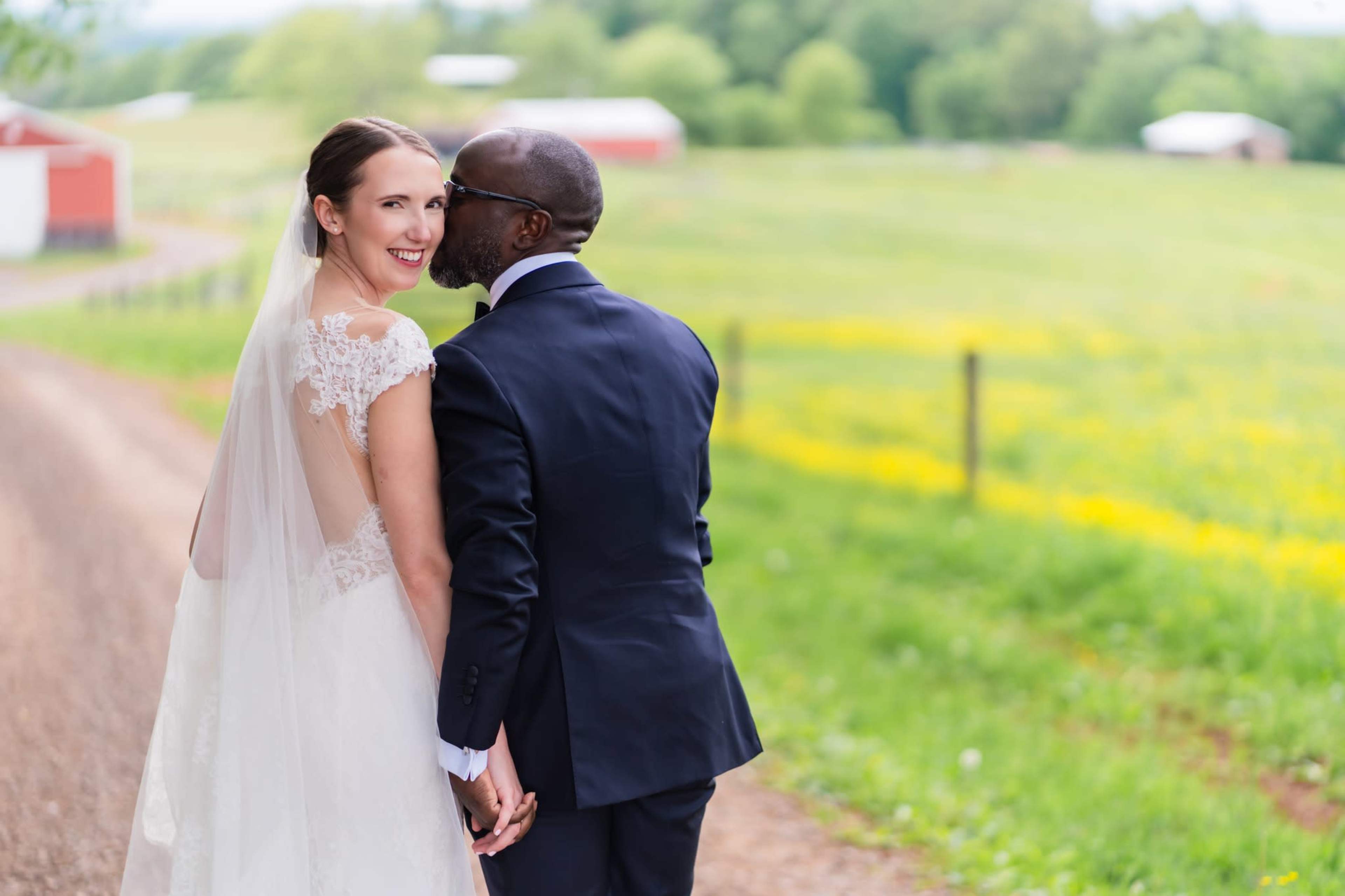 A bride and groom hold hands and gaze at each other while standing on a dirt road in a rural setting.