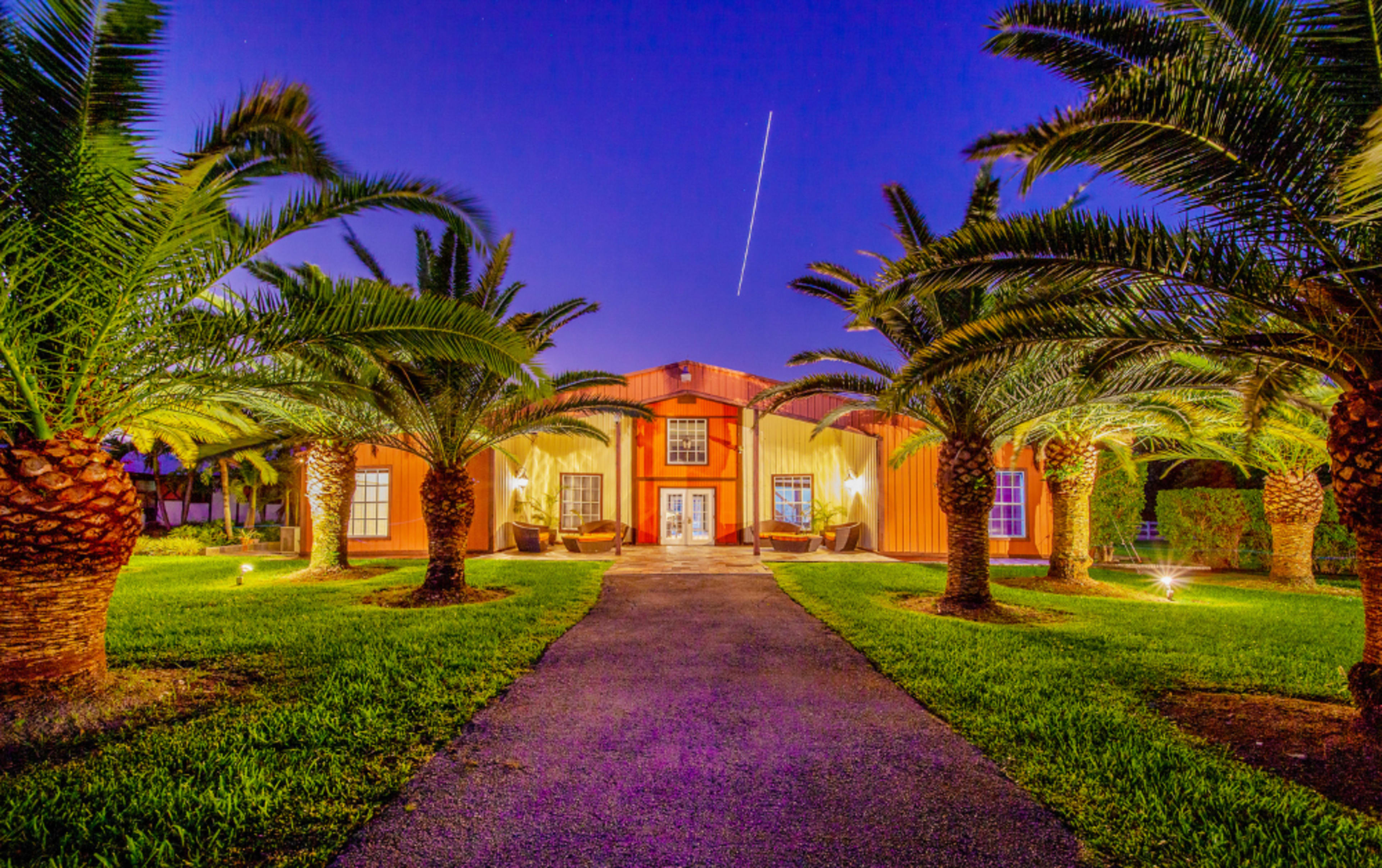 A pathway lined with palm trees leads to a two-story orange building under a twilight sky.