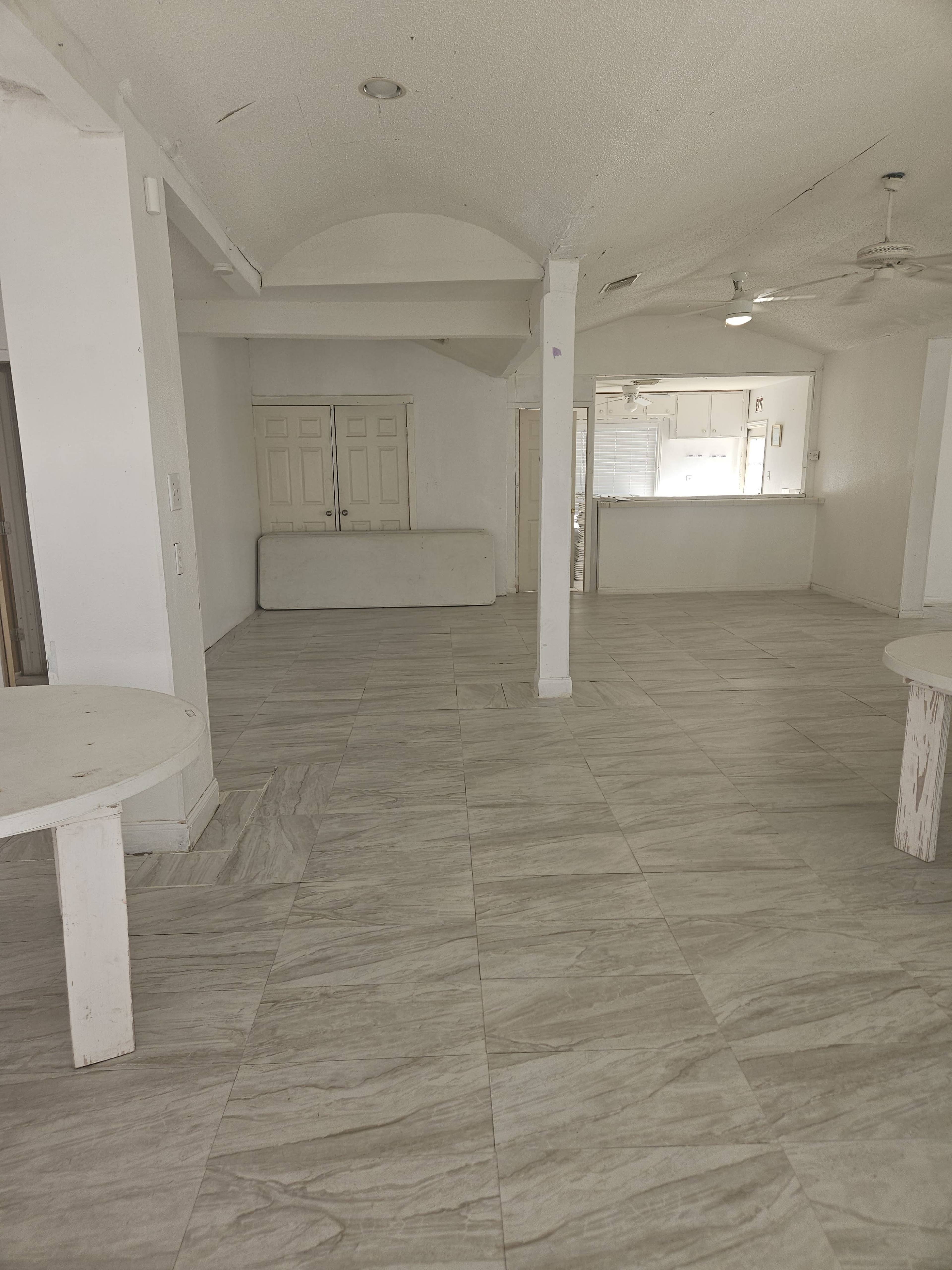 The image shows a spacious interior of a room with tiled flooring, two white circular tables, and a kitchenette area in the background.