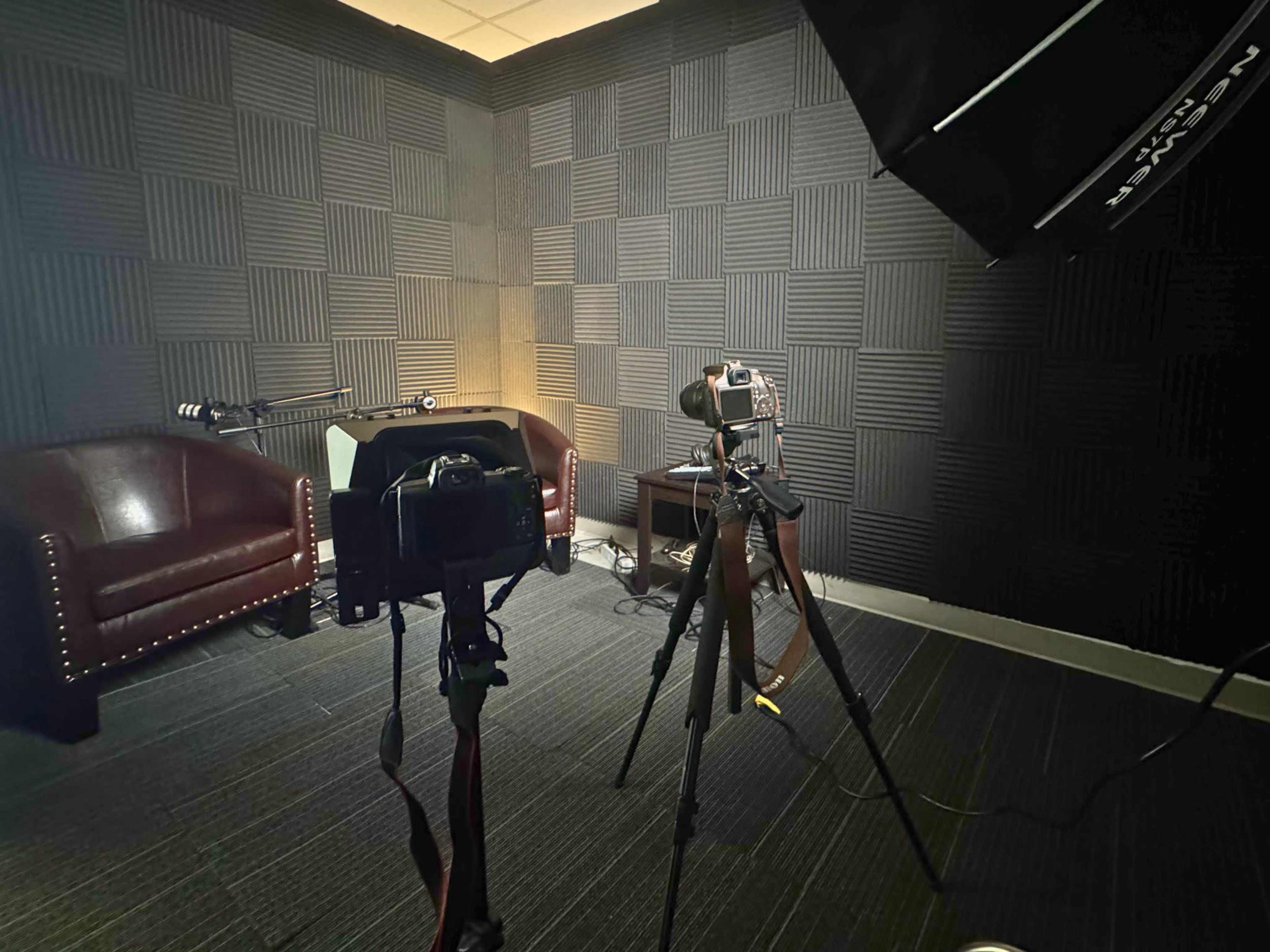 The image shows a small recording studio with two leather chairs, a camera mounted on a tripod, and soft lighting equipment.