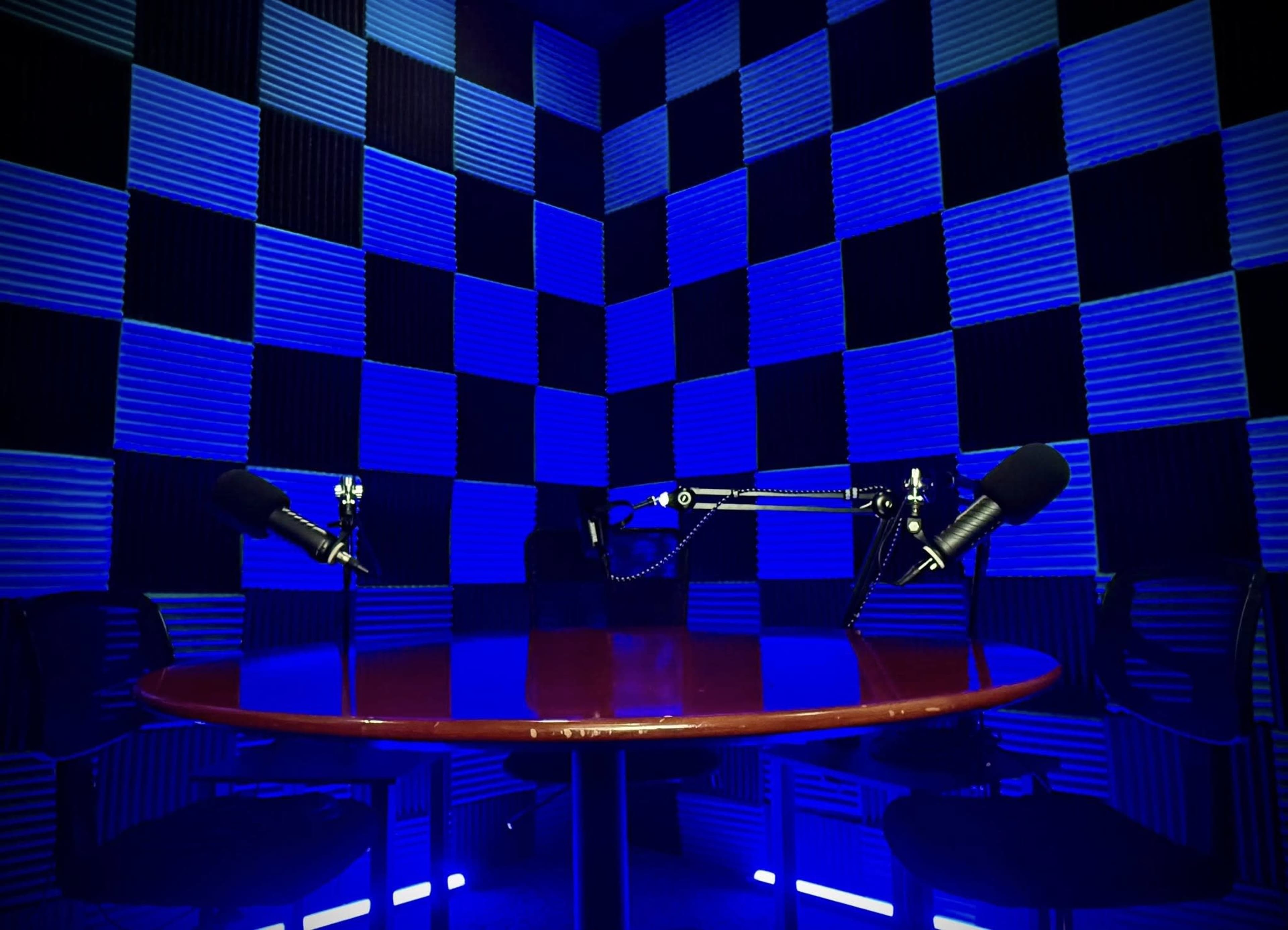 The image shows a podcast recording studio with a round table and two microphones set against a backdrop of blue and black checkered walls.