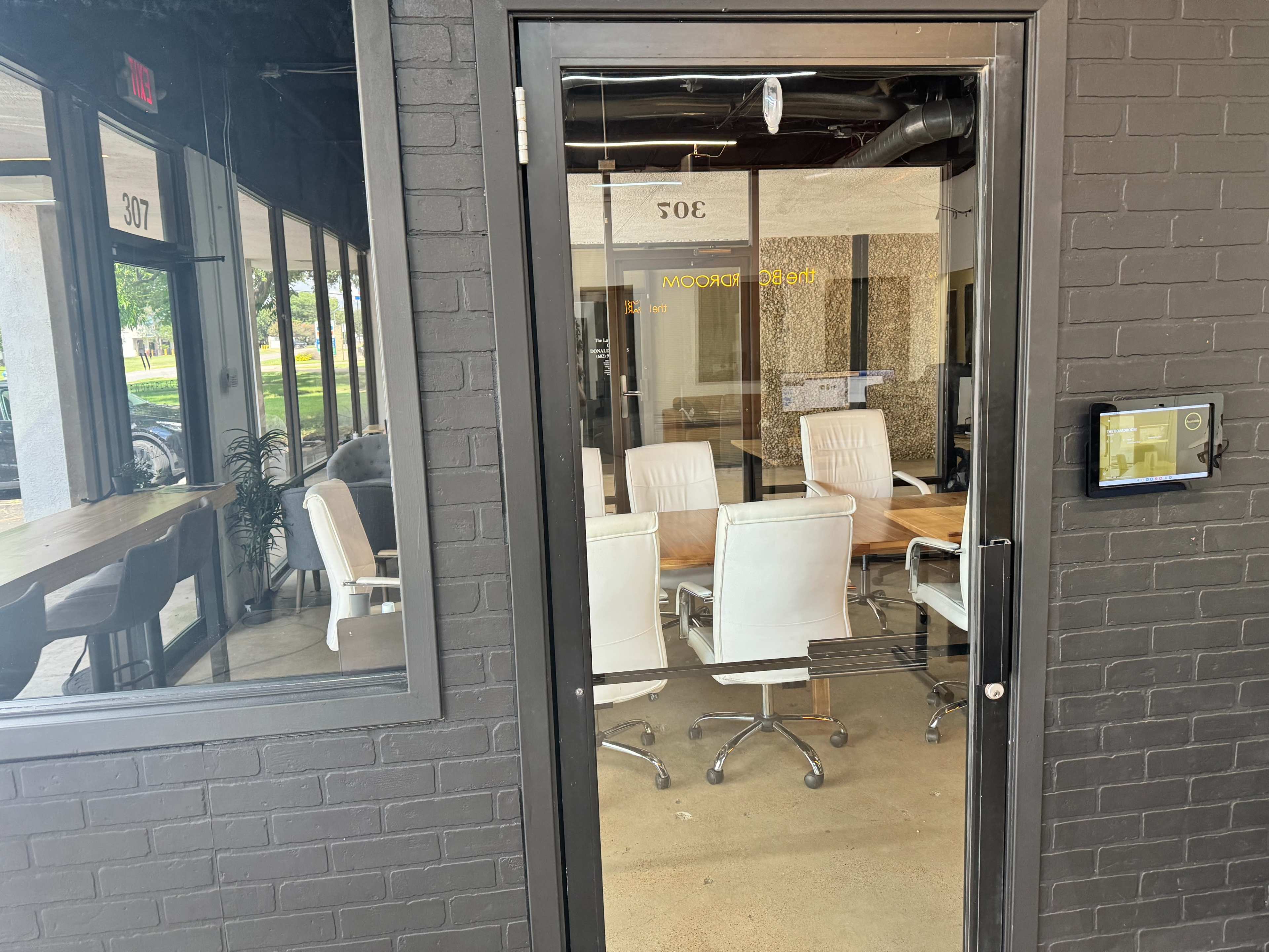 The image shows a glass door leading into a modern conference room with a wooden table and white chairs.