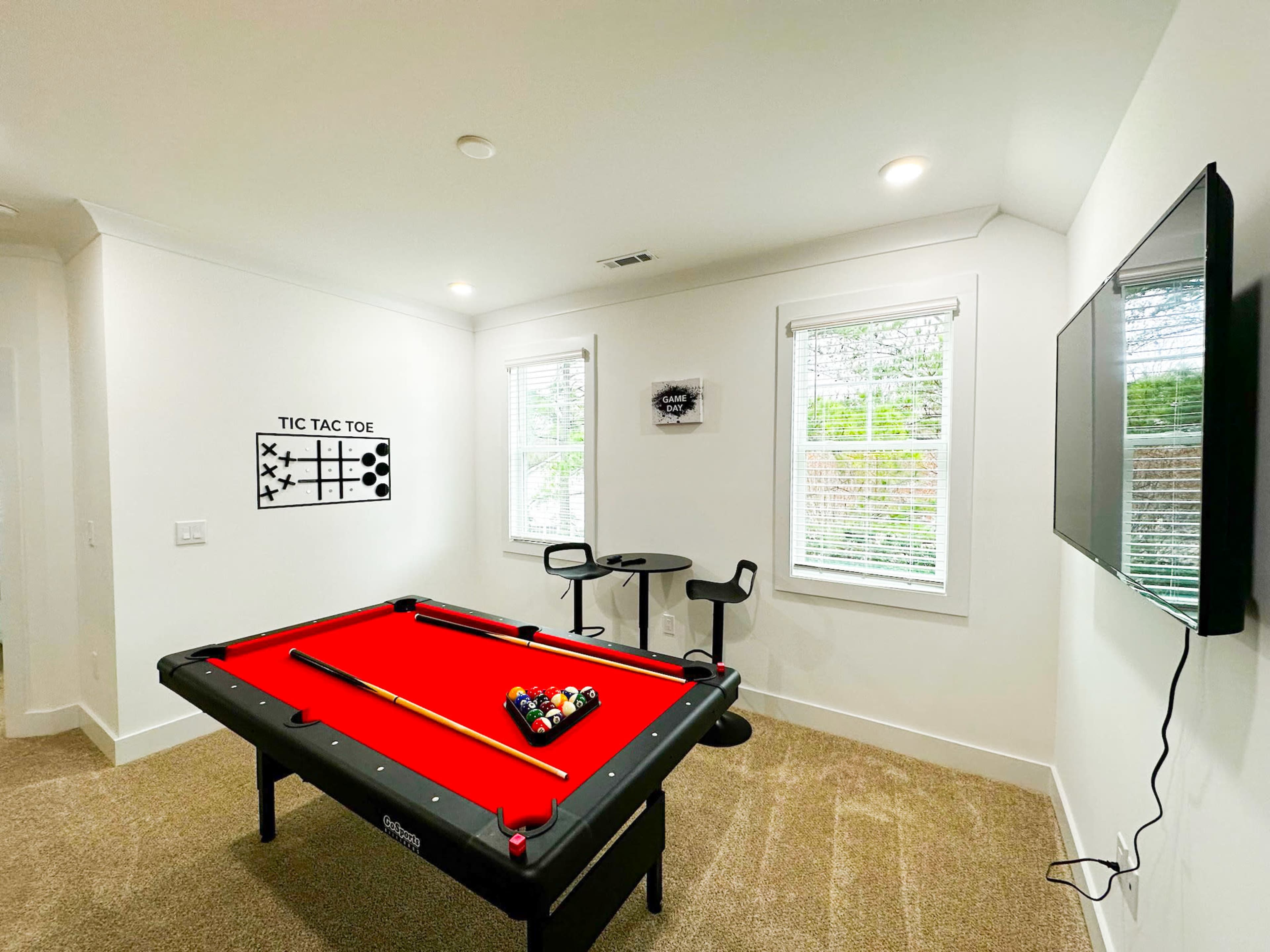 The image shows a game room featuring a red pool table, a small table with two chairs, a tic-tac-toe wall design, and a TV mounted on the wall.