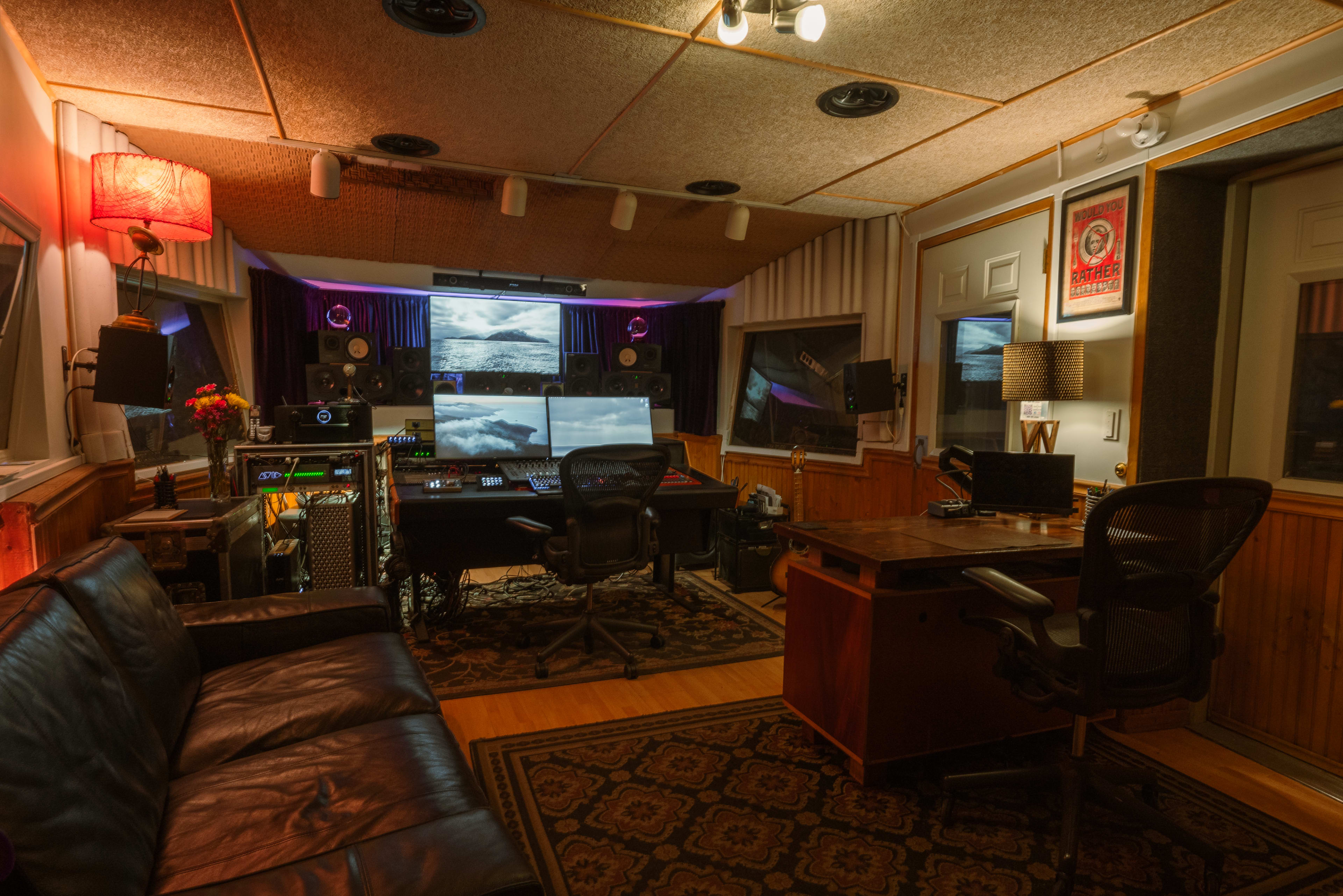 The image shows a dimly lit recording studio with a large sound mixing console, two computer desks, and a sofa, adorned with decorative lamps and wall art.