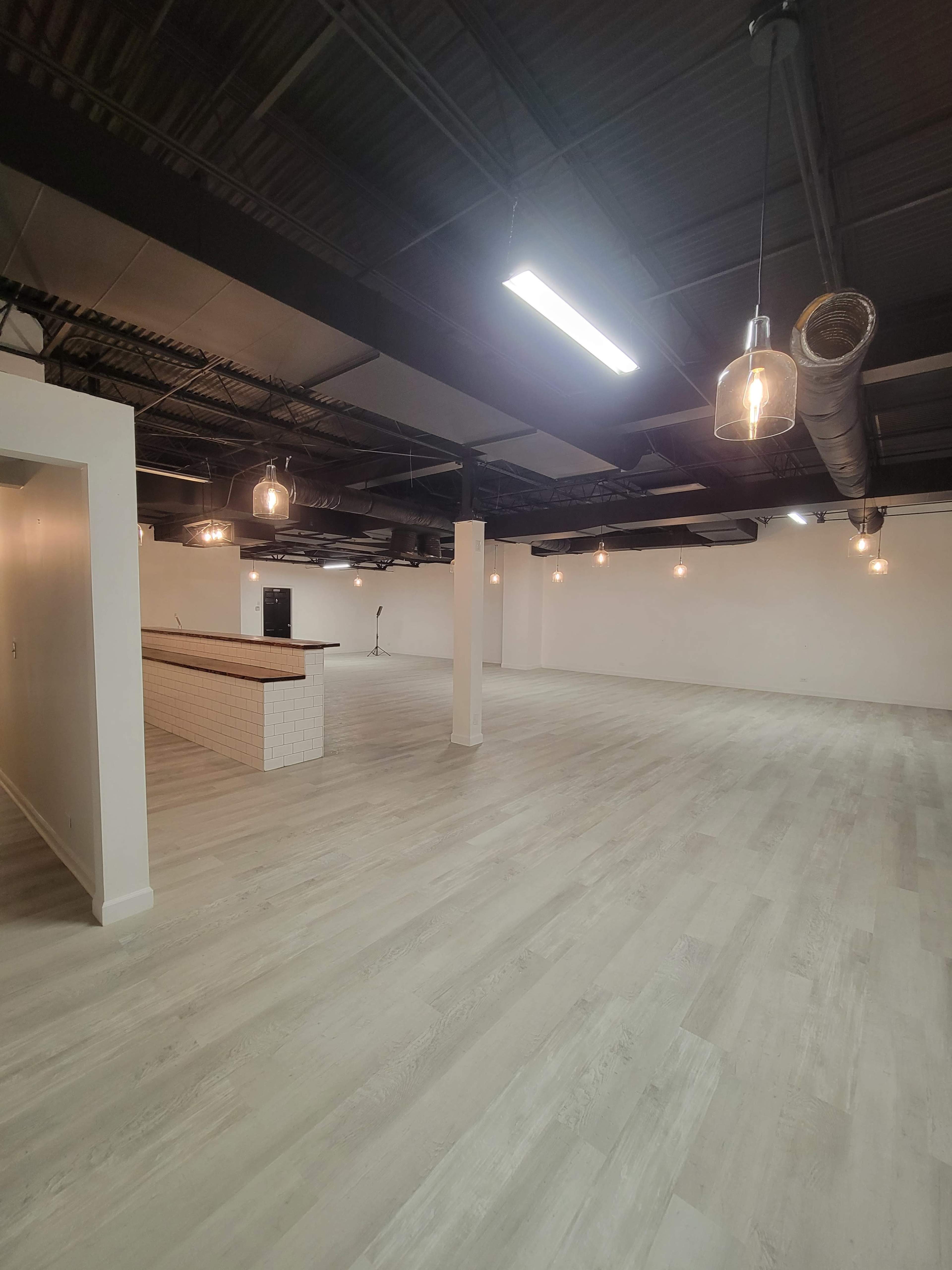An empty commercial space with light-colored flooring, exposed ductwork, and pendant lights hanging from the ceiling.
