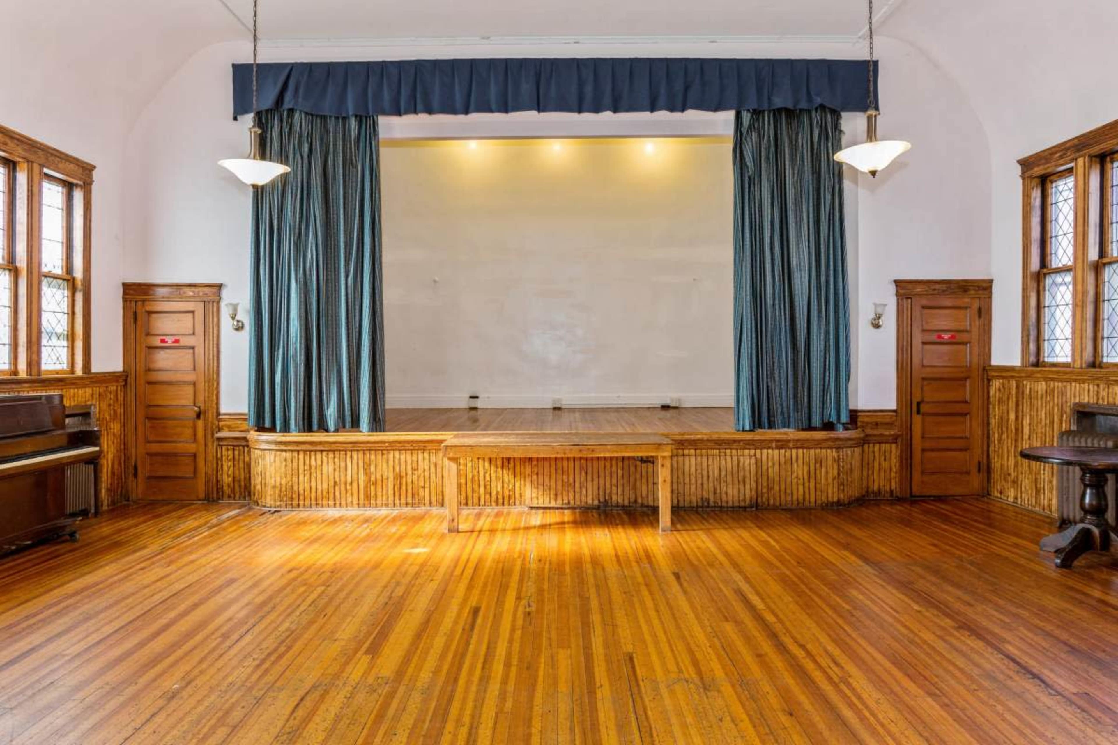 A well-lit performance stage with wooden flooring, flanked by dark blue curtains and wooden-paneled walls with doors on either side.